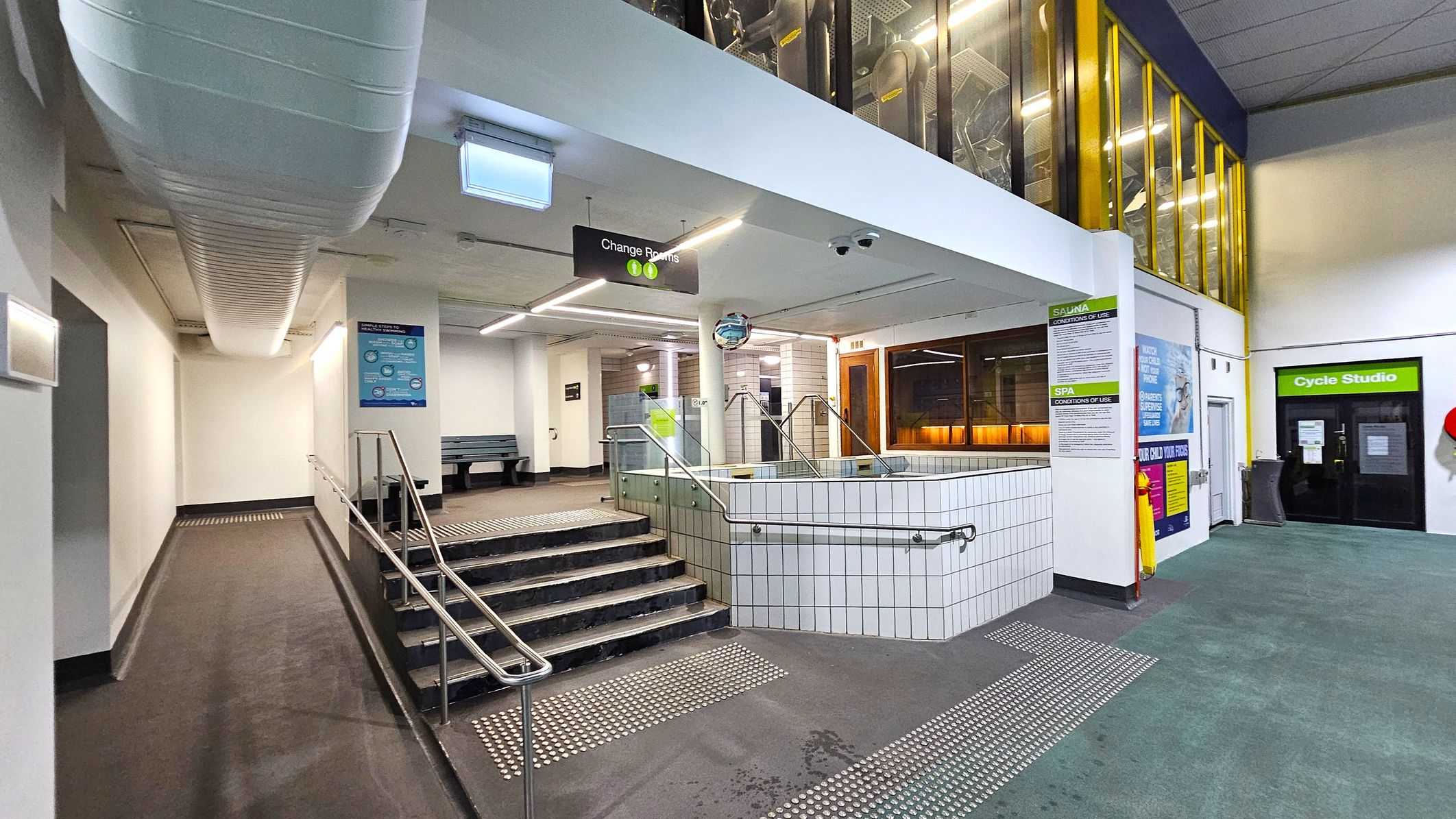 A modern indoor facility with a tiled hot tub in the centre, surrounded by metal railings. To the left, a ramp and stairs lead to a corridor with a bench. Overhead, a large white duct runs along the ceiling. On the right, a glass-walled area with yellow frames and a door labelled "Cycle Studio". Signs for changing rooms and spa conditions are visible. The floor is a mix of textured tiles and carpet, and the ceiling is fitted with fluorescent lights. A modern indoor facility with a tiled hot tub in the centre, surrounded by metal railings. To the left, a ramp and stairs lead to a corridor with a bench. Overhead, a large white duct runs along the ceiling. On the right, a glass-walled area with yellow frames and a door labelled "Cycle Studio". Signs for changing rooms and spa conditions are visible. The floor is a mix of textured tiles and carpet, and the ceiling is fitted with fluorescent lights.