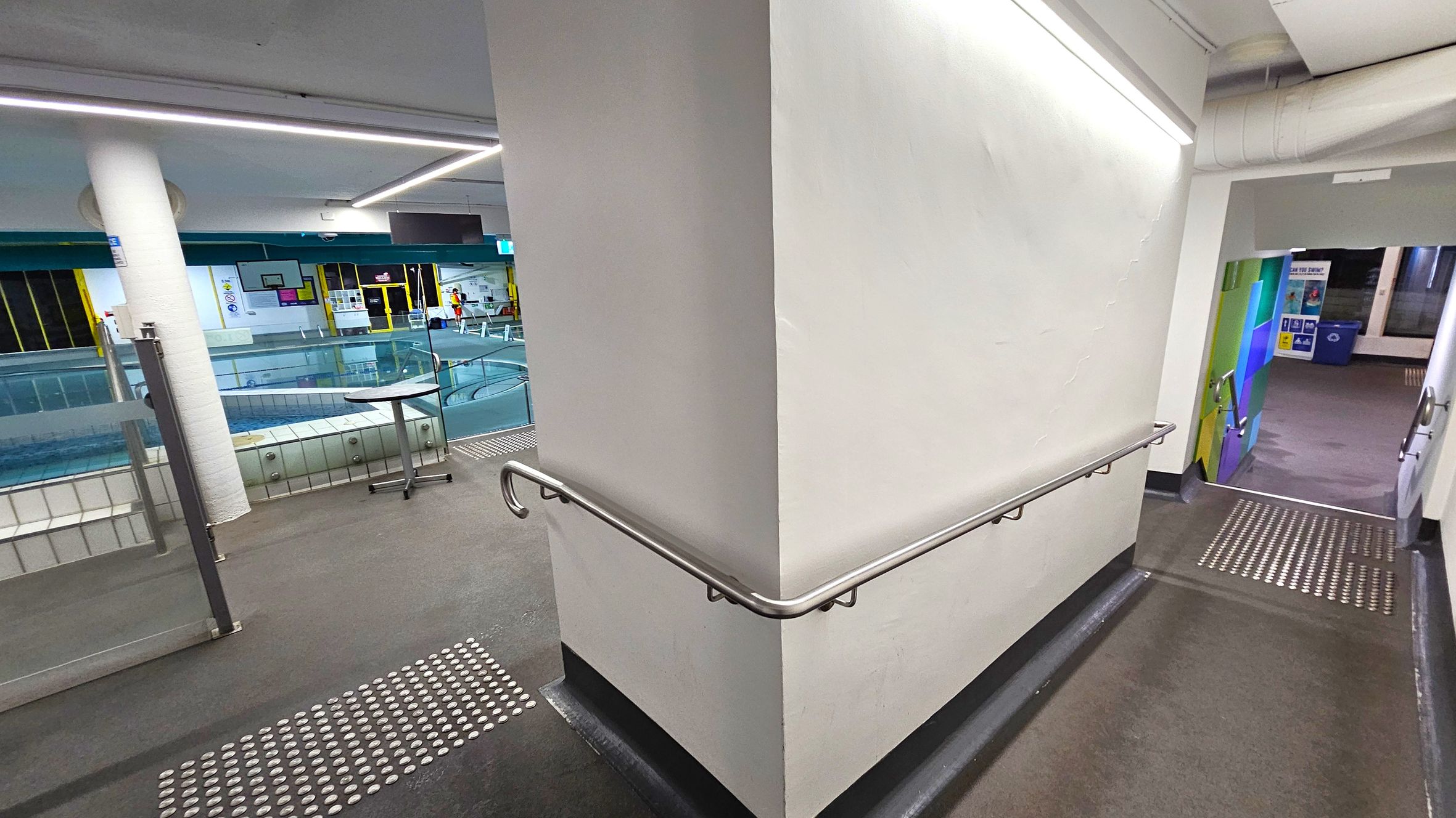 A corridor with a white wall in the centre, featuring a metal handrail. On the left, there's a view of an indoor swimming pool with glass barriers and a small round table. The pool area has yellow-framed windows and various signs. On the right, a ramp with tactile paving leads downwards, with colourful wall panels visible at the bottom. The ceiling has linear lighting, and the floor is dark with tactile indicators. A corridor with a white wall in the centre, featuring a metal handrail. On the left, there's a view of an indoor swimming pool with glass barriers and a small round table. The pool area has yellow-framed windows and various signs. On the right, a ramp with tactile paving leads downwards, with colourful wall panels visible at the bottom. The ceiling has linear lighting, and the floor is dark with tactile indicators.