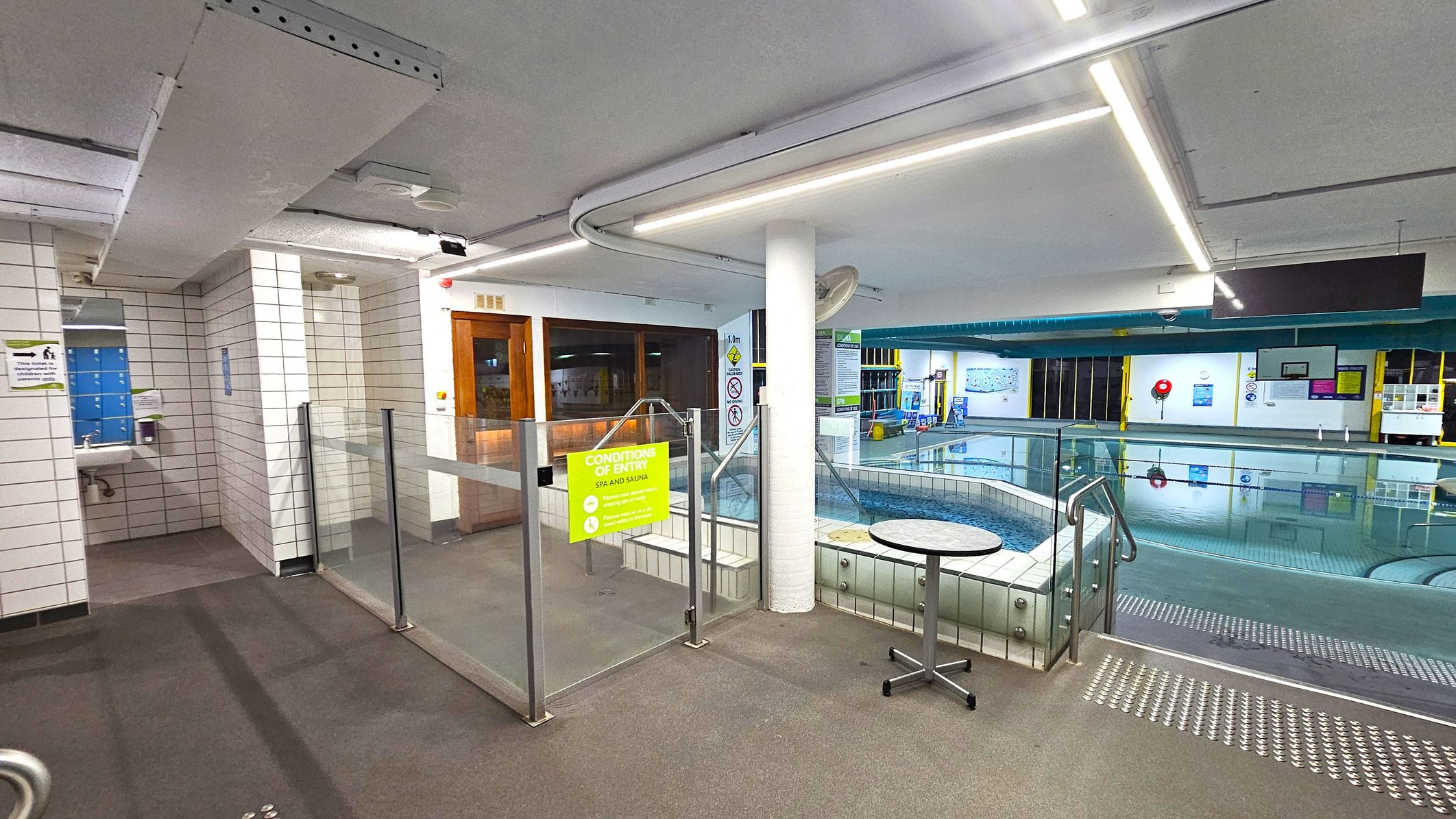 A swimming pool area with a tiled floor and ceiling. On the left, there is a sink and mirror next to a tiled wall. In the centre, a glass barrier with a yellow sign reading "Conditions of Entry" separates the pool area. A small round table is nearby. On the right, the pool is visible with clear water, surrounded by more tiled surfaces. The background shows various signs and equipment, with a lifebuoy on the wall. The ceiling has fluorescent lighting. A swimming pool area with a tiled floor and ceiling. On the left, there is a sink and mirror next to a tiled wall. In the centre, a glass barrier with a yellow sign reading "Conditions of Entry" separates the pool area. A small round table is nearby. On the right, the pool is visible with clear water, surrounded by more tiled surfaces. The background shows various signs and equipment, with a lifebuoy on the wall. The ceiling has fluorescent lighting.