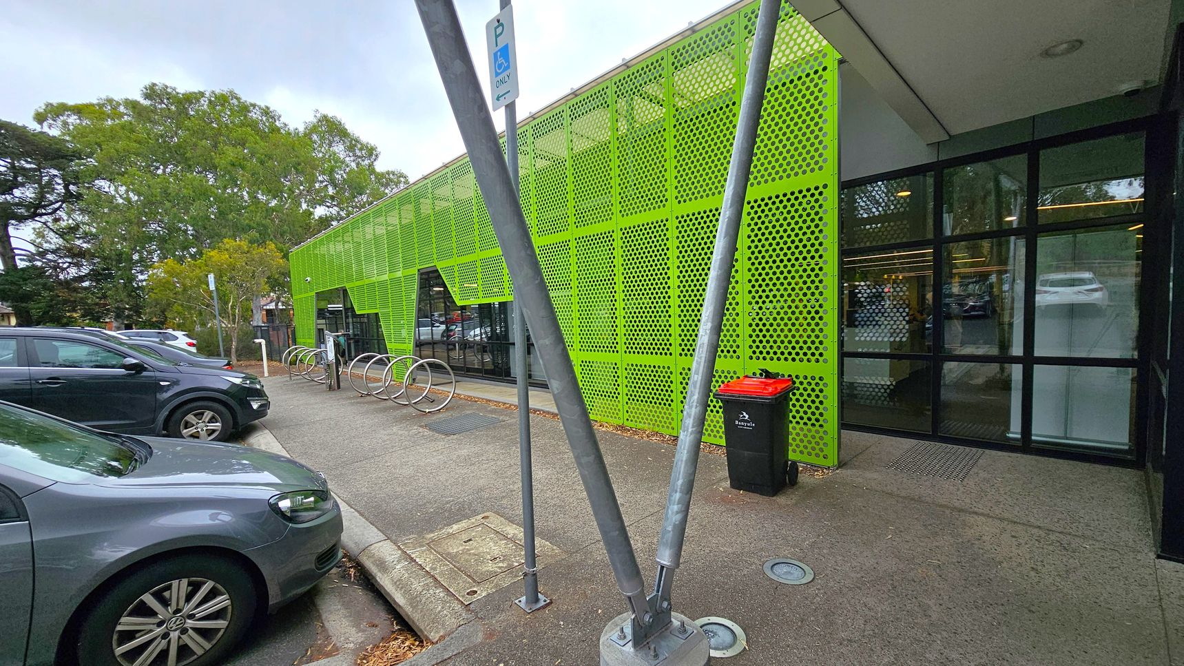 First sequence green facade. Side Entrance to Studios. Ivanhoe Aquatic Centre. A modern building with a bright green perforated metal facade on the right. Large glass windows are visible beneath the green panels. In front, there are metal bike racks and a black bin with a red lid. Two metal poles are positioned in the foreground. On the left, several parked cars are visible, and trees are in the background. The ground is paved, and the ceiling of the building is flat and white. First sequence green facade. Side Entrance to Studios. Ivanhoe Aquatic Centre. A modern building with a bright green perforated metal facade on the right. Large glass windows are visible beneath the green panels. In front, there are metal bike racks and a black bin with a red lid. Two metal poles are positioned in the foreground. On the left, several parked cars are visible, and trees are in the background. The ground is paved, and the ceiling of the building is flat and white.