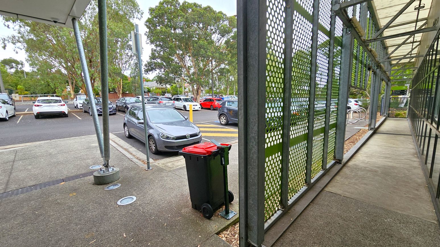 Second sequence sloping pathway. Side Entrance to Studios. Ivanhoe Aquatic Centre. A parking area with several cars is visible on the left, surrounded by trees. A grey car is parked near a red-lidded rubbish bin on a paved surface. On the right, a walkway is enclosed by a metal structure with perforated panels and a transparent roof. The ground is a mix of concrete and green patches. Second sequence sloping pathway. Side Entrance to Studios. Ivanhoe Aquatic Centre. A parking area with several cars is visible on the left, surrounded by trees. A grey car is parked near a red-lidded rubbish bin on a paved surface. On the right, a walkway is enclosed by a metal structure with perforated panels and a transparent roof. The ground is a mix of concrete and green patches.