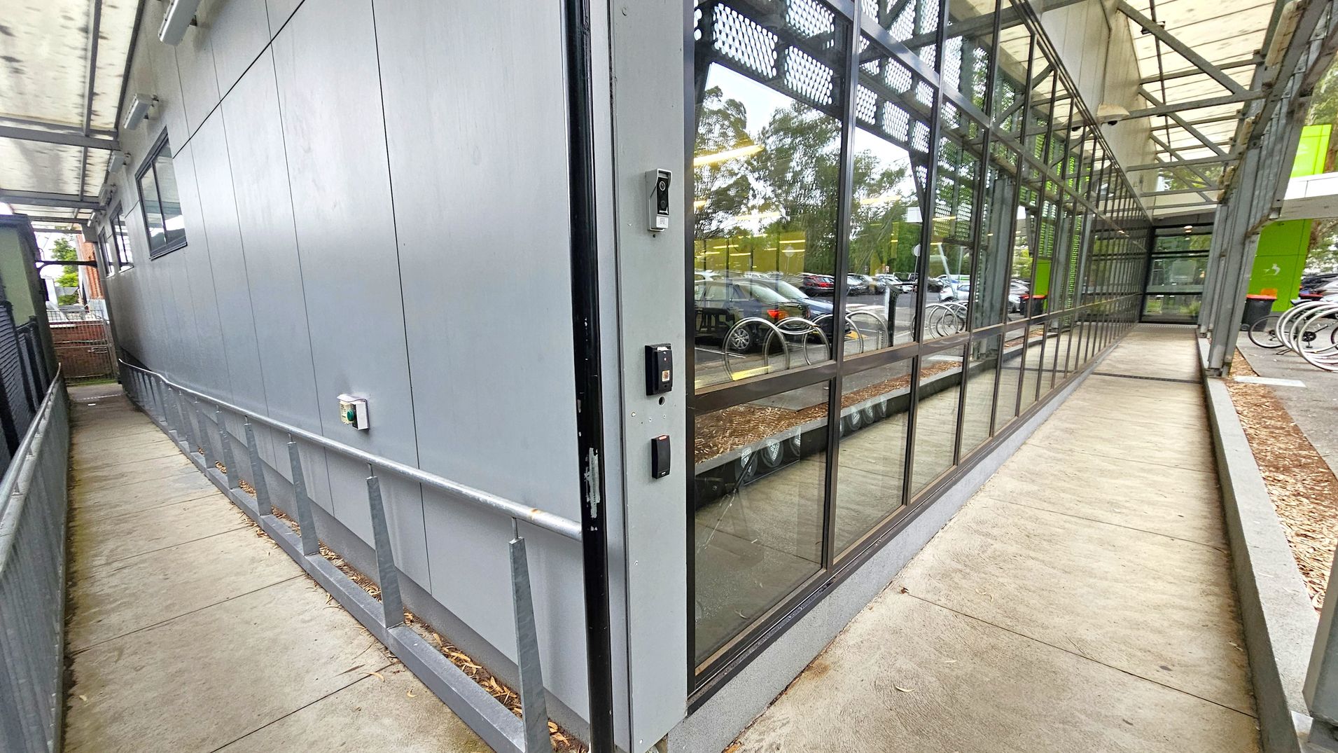 Third sequence gated corridor. Side Entrance to Studios. Ivanhoe Aquatic Centre. A narrow walkway with a metal railing on the left, adjacent to a grey building wall with a small window. On the right, a glass wall reflects parked cars and trees. The path is covered by a transparent roof supported by metal beams. The floor is concrete, and there are security devices mounted on the wall near the glass section. In the background, bicycles are parked near a green structure. Third sequence gated corridor. Side Entrance to Studios. Ivanhoe Aquatic Centre. A narrow walkway with a metal railing on the left, adjacent to a grey building wall with a small window. On the right, a glass wall reflects parked cars and trees. The path is covered by a transparent roof supported by metal beams. The floor is concrete, and there are security devices mounted on the wall near the glass section. In the background, bicycles are parked near a green structure.