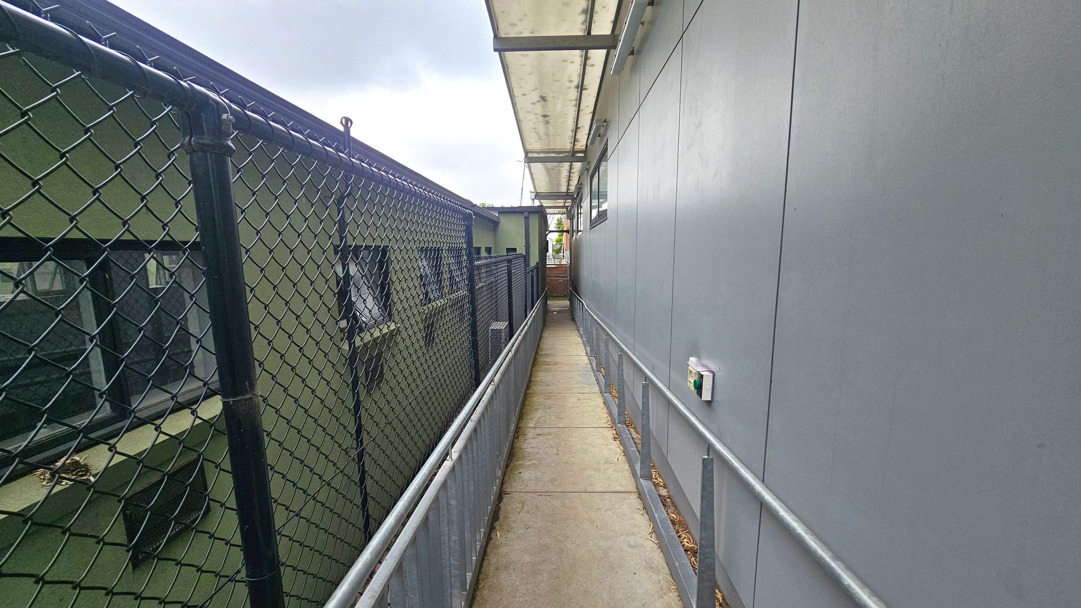 Fourth sequence narrow walkway. Side Entrance to Studios. Ivanhoe Aquatic Centre. A narrow walkway with a concrete floor runs between two structures. On the left, there's a green building with windows, separated by a chain-link fence. On the right, a grey building with a smooth wall and a small device attached. Above, a translucent canopy provides cover. The walkway is bordered by metal railings on both sides. Fourth sequence narrow walkway. Side Entrance to Studios. Ivanhoe Aquatic Centre. A narrow walkway with a concrete floor runs between two structures. On the left, there's a green building with windows, separated by a chain-link fence. On the right, a grey building with a smooth wall and a small device attached. Above, a translucent canopy provides cover. The walkway is bordered by metal railings on both sides.