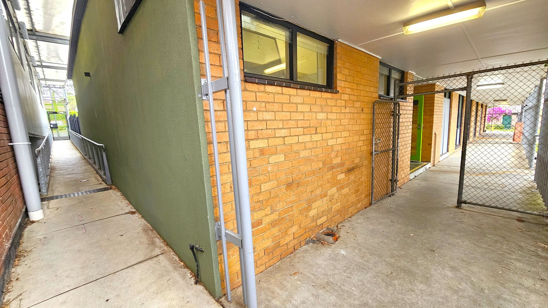 Fifth sequence corridor to studios. Side Entrance to Studios. Ivanhoe Aquatic Centre. A narrow passageway with a concrete floor. On the left, a green wall with a metal railing leads to a gate. On the right, a brick wall with windows above and a chain-link gate. The ceiling is white with fluorescent lights. In the background, a corridor with more brick walls and a glimpse of greenery and a purple flowering bush. Fifth sequence corridor to studios. Side Entrance to Studios. Ivanhoe Aquatic Centre. A narrow passageway with a concrete floor. On the left, a green wall with a metal railing leads to a gate. On the right, a brick wall with windows above and a chain-link gate. The ceiling is white with fluorescent lights. In the background, a corridor with more brick walls and a glimpse of greenery and a purple flowering bush.