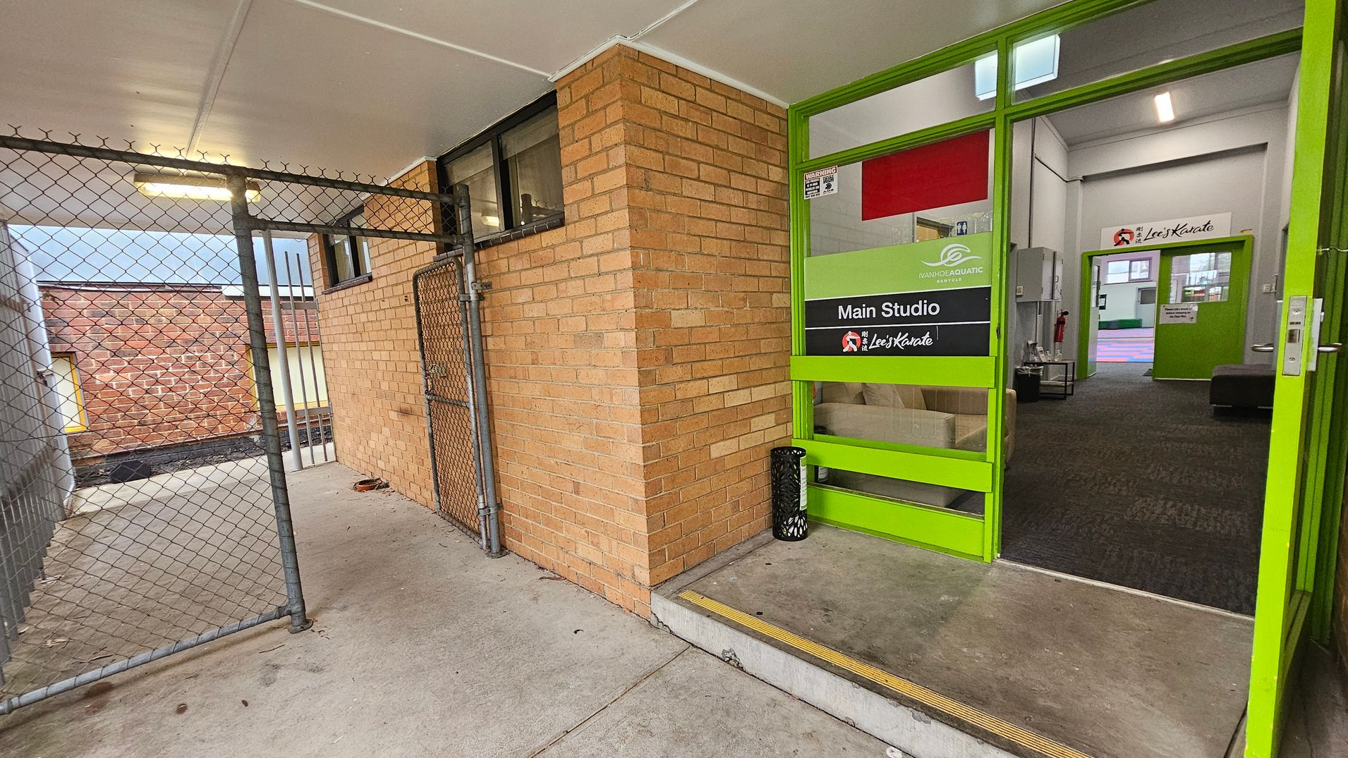 A corridor with a chain-link fence on the left and a brick wall on the right. The floor is concrete. The entrance to a building is on the right, featuring a green-framed glass door with signs for "Main Studio" and "Lee's Karate." Inside, the carpeted floor is visible, along with a fire extinguisher and a small table. The ceiling is white with fluorescent lighting. A corridor with a chain-link fence on the left and a brick wall on the right. The floor is concrete. The entrance to a building is on the right, featuring a green-framed glass door with signs for "Main Studio" and "Lee's Karate." Inside, the carpeted floor is visible, along with a fire extinguisher and a small table. The ceiling is white with fluorescent lighting.