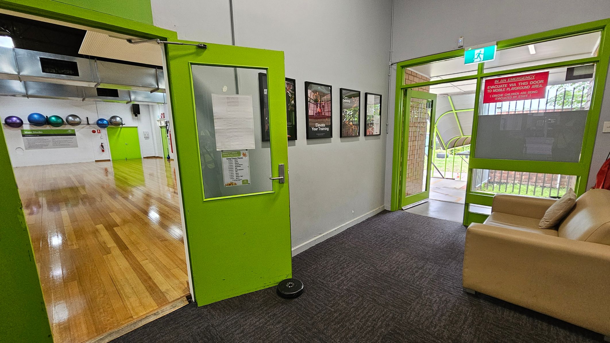 A room with bright green doors and a grey carpeted floor. On the left, an open door leads to a wooden-floored studio with exercise balls on the wall. On the right, another green-framed door opens to an outdoor area with grass visible. A beige sofa with a cushion is in the foreground. Several framed posters are mounted on the wall between the doors. An emergency exit sign is above the right door. A room with bright green doors and a grey carpeted floor. On the left, an open door leads to a wooden-floored studio with exercise balls on the wall. On the right, another green-framed door opens to an outdoor area with grass visible. A beige sofa with a cushion is in the foreground. Several framed posters are mounted on the wall between the doors. An emergency exit sign is above the right door.