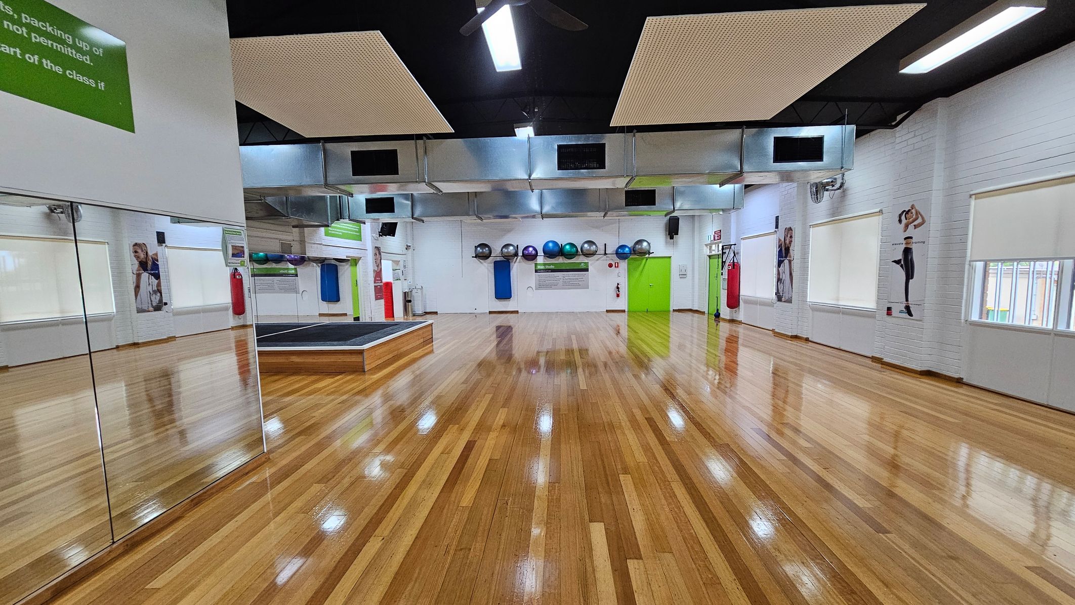 A spacious gym room with polished wooden floors. On the left, there are large mirrors reflecting the room. The back wall features exercise balls in various colours on a rack, a green door, and a small stage. The right wall has posters of people exercising and several windows with blinds. The ceiling is black with rectangular light fixtures and ventilation ducts. A red punching bag hangs near the back wall. A spacious gym room with polished wooden floors. On the left, there are large mirrors reflecting the room. The back wall features exercise balls in various colours on a rack, a green door, and a small stage. The right wall has posters of people exercising and several windows with blinds. The ceiling is black with rectangular light fixtures and ventilation ducts. A red punching bag hangs near the back wall.