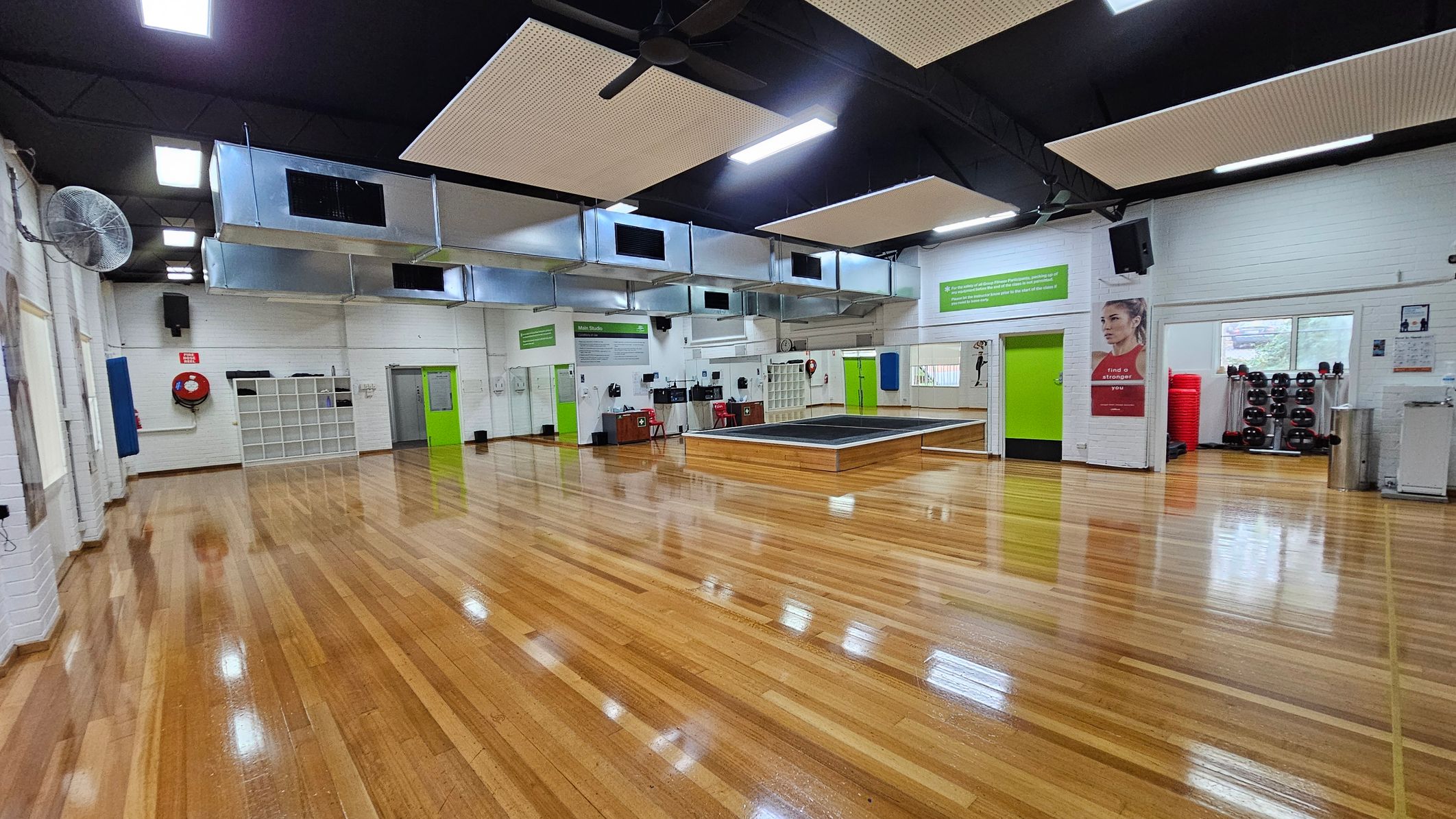 A spacious gym studio with polished wooden floors and a high ceiling featuring large ventilation ducts and ceiling fans. On the left, there are white brick walls with a fire hose reel and shelving units. In the centre, a raised platform is visible. On the right, a green door is flanked by posters and a window. Exercise equipment, including weights and stacked mats, is neatly arranged. Bright overhead lights illuminate the room. A spacious gym studio with polished wooden floors and a high ceiling featuring large ventilation ducts and ceiling fans. On the left, there are white brick walls with a fire hose reel and shelving units. In the centre, a raised platform is visible. On the right, a green door is flanked by posters and a window. Exercise equipment, including weights and stacked mats, is neatly arranged. Bright overhead lights illuminate the room.