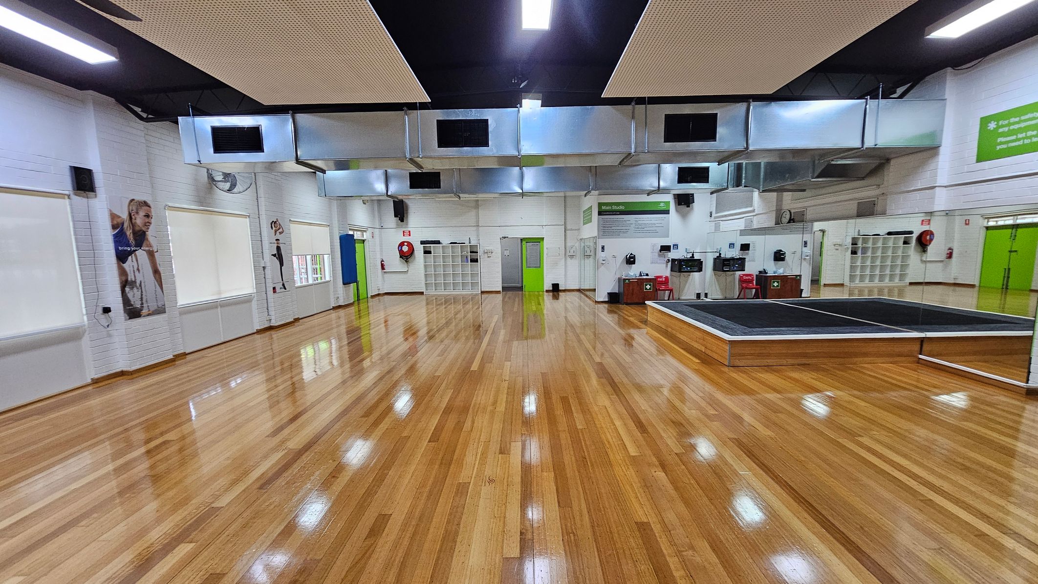 A spacious room with polished wooden floors and white brick walls. On the left, there are large windows with blinds and posters of athletes. The centre features a green door and a wall with cubbyholes. On the right, there's a raised platform with mirrors and a green sign above. The ceiling has large metallic air ducts and rectangular lights. Red chairs and fire safety equipment are visible along the walls. A spacious room with polished wooden floors and white brick walls. On the left, there are large windows with blinds and posters of athletes. The centre features a green door and a wall with cubbyholes. On the right, there's a raised platform with mirrors and a green sign above. The ceiling has large metallic air ducts and rectangular lights. Red chairs and fire safety equipment are visible along the walls.