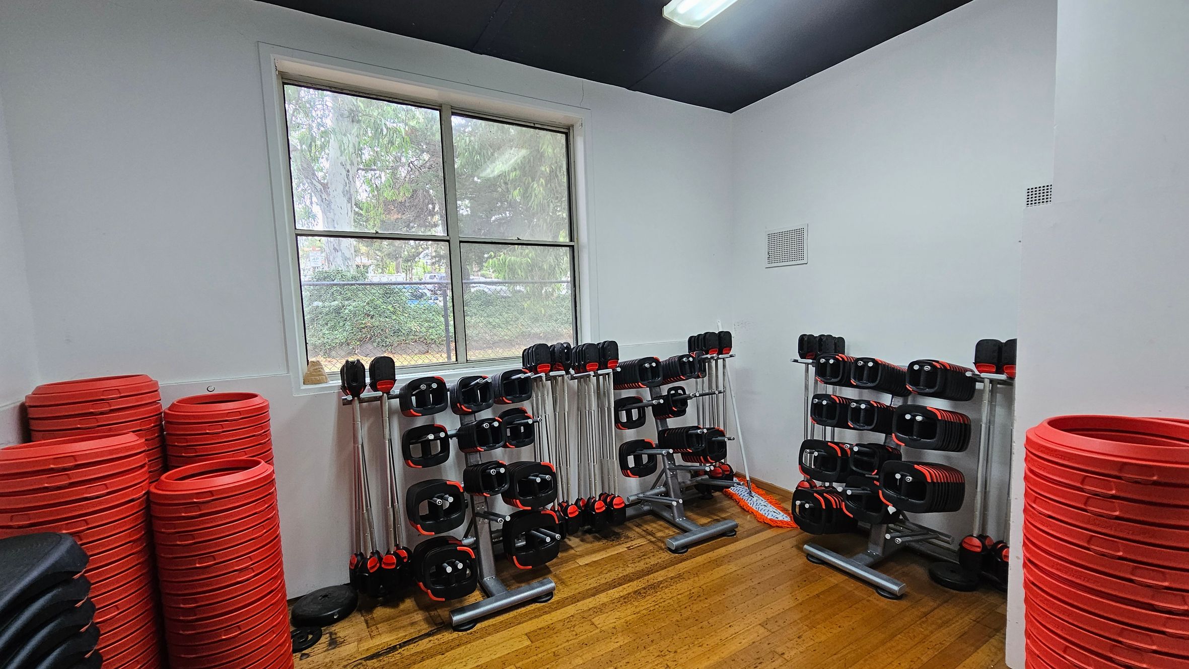 A gym room with wooden flooring and a black ceiling. On the left, there are stacks of red weight plates and black step platforms. In the centre, several racks hold barbells with black and red weights. A window in the background shows greenery outside. On the right, more red weight plates are stacked. A mop is leaning against the wall near the racks. A gym room with wooden flooring and a black ceiling. On the left, there are stacks of red weight plates and black step platforms. In the centre, several racks hold barbells with black and red weights. A window in the background shows greenery outside. On the right, more red weight plates are stacked. A mop is leaning against the wall near the racks.