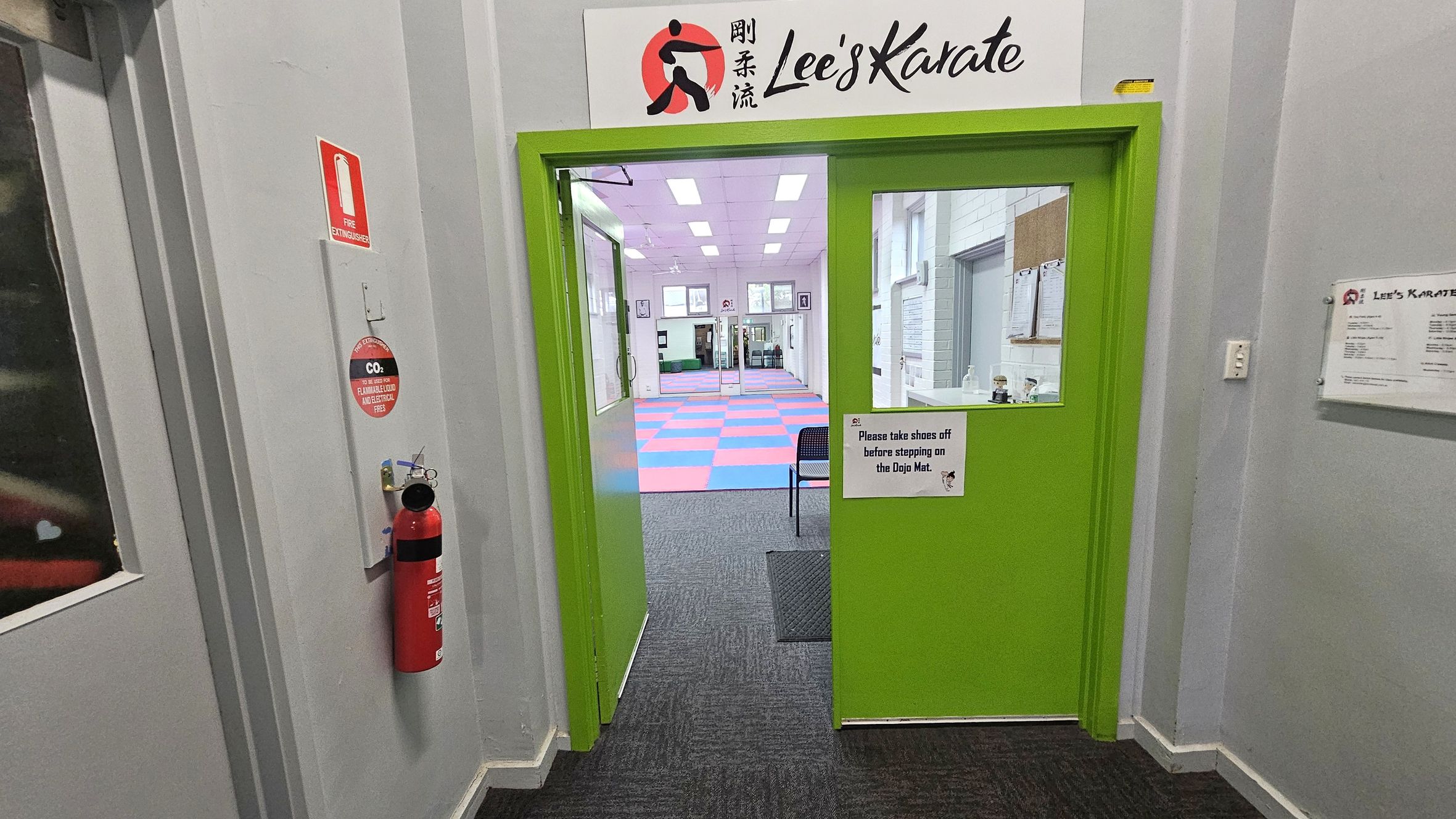 A bright green door with a sign above it reading "Lee's Karate" leads into a room with a colourful checkered mat. A sign on the door requests visitors to remove shoes before stepping on the mat. To the left, a red fire extinguisher is mounted on the wall. The floor is carpeted in dark grey, and the walls are light grey. Inside the room, there are framed pictures and a chair visible. The ceiling has rectangular lights. A bright green door with a sign above it reading "Lee's Karate" leads into a room with a colourful checkered mat. A sign on the door requests visitors to remove shoes before stepping on the mat. To the left, a red fire extinguisher is mounted on the wall. The floor is carpeted in dark grey, and the walls are light grey. Inside the room, there are framed pictures and a chair visible. The ceiling has rectangular lights.