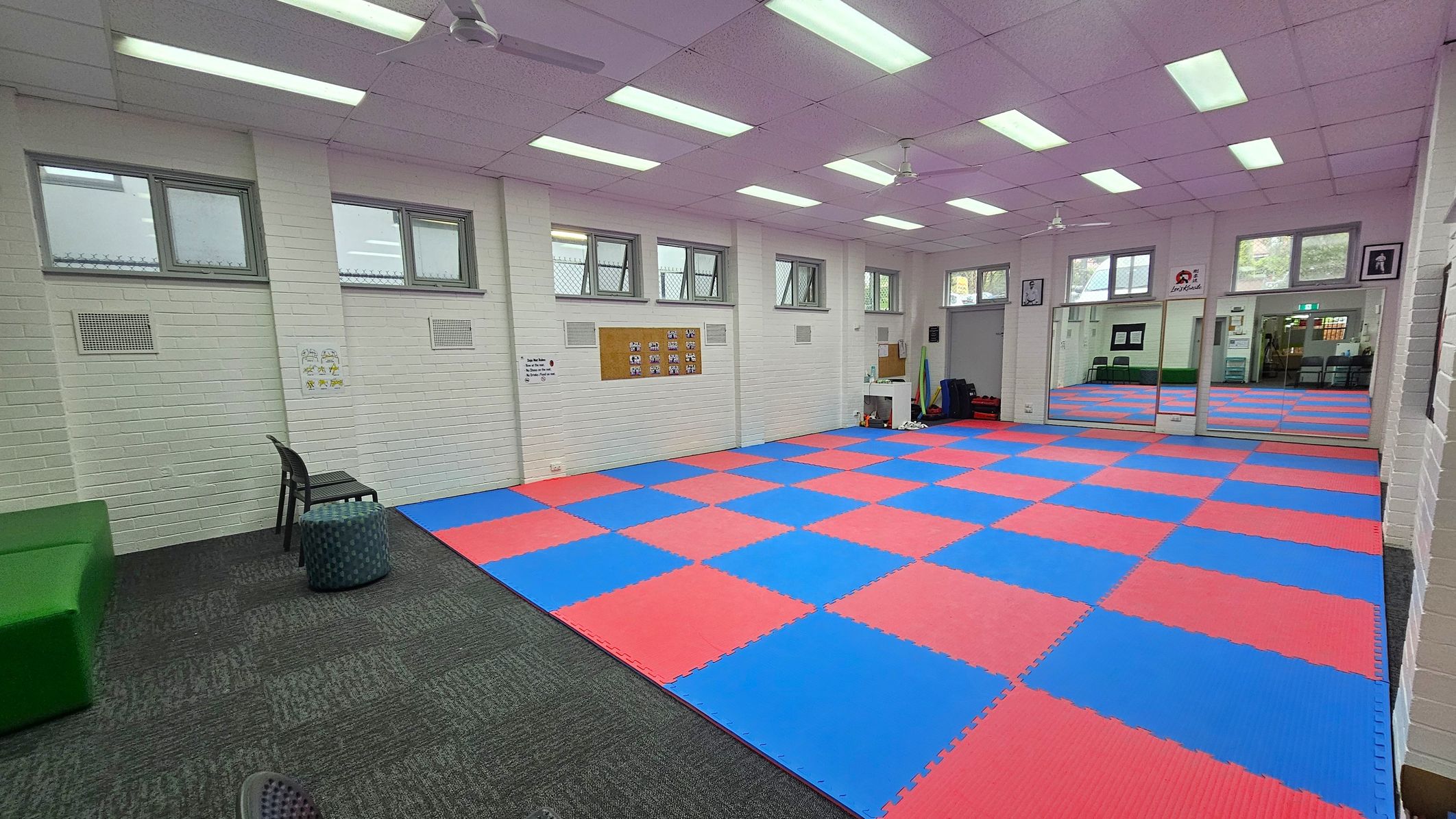 A room with a blue and red checkered mat covering most of the floor. On the left, there is a green bench, a black chair, and a small round stool on a grey carpeted area. The walls are white with several small windows near the ceiling. On the right, there are large mirrors and a doorway leading to another room. The ceiling has fluorescent lights and ceiling fans. Various posters and a bulletin board are on the walls. A room with a blue and red checkered mat covering most of the floor. On the left, there is a green bench, a black chair, and a small round stool on a grey carpeted area. The walls are white with several small windows near the ceiling. On the right, there are large mirrors and a doorway leading to another room. The ceiling has fluorescent lights and ceiling fans. Various posters and a bulletin board are on the walls.