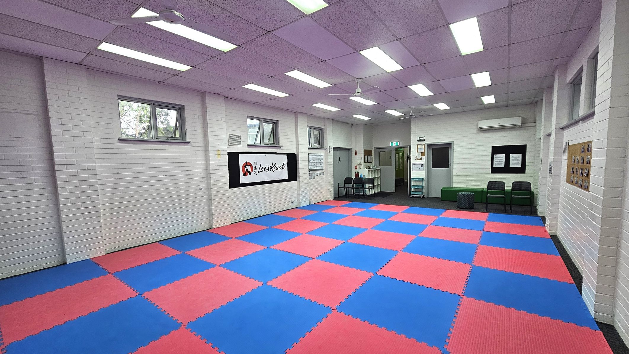 A martial arts studio with a floor covered in interlocking red and blue mats. The walls are white brick with several small windows near the ceiling. On the left wall, there is a sign with text and a logo. The right side has a bulletin board with photos. At the back, there are chairs, a green bench, and a small table. The ceiling is purple with fluorescent lights and a ceiling fan. A martial arts studio with a floor covered in interlocking red and blue mats. The walls are white brick with several small windows near the ceiling. On the left wall, there is a sign with text and a logo. The right side has a bulletin board with photos. At the back, there are chairs, a green bench, and a small table. The ceiling is purple with fluorescent lights and a ceiling fan.