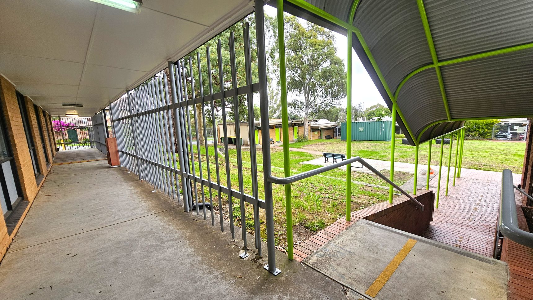 Covered walkway. Functional and Barre Studio. Ivanhoe Aquatic Centre. A covered walkway with a concrete floor and a white ceiling on the left, featuring brick walls and windows. Metal bars form a fence along the side. On the right, a ramp with a handrail leads down to a brick path. The roof above the ramp is supported by green poles. In the background, there is a grassy area with trees, a green shipping container, and a few small buildings. A bench is visible on the grass. Covered walkway. Functional and Barre Studio. Ivanhoe Aquatic Centre. A covered walkway with a concrete floor and a white ceiling on the left, featuring brick walls and windows. Metal bars form a fence along the side. On the right, a ramp with a handrail leads down to a brick path. The roof above the ramp is supported by green poles. In the background, there is a grassy area with trees, a green shipping container, and a few small buildings. A bench is visible on the grass.