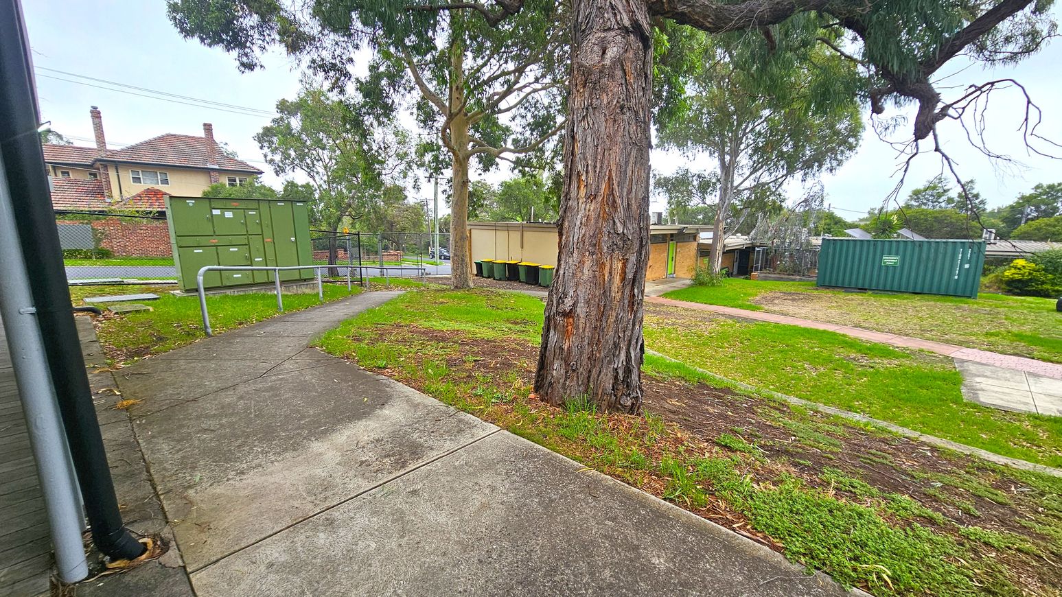Concrete walkways. Functional and Barre Studio. Ivanhoe Aquatic Centre. A concrete path curves through a grassy area with a large tree in the centre. On the left, there is a green utility box and a brick building with a tiled roof in the background. On the right, a green shipping container and several wheelie bins are visible near a small building. Trees and a fence line the background, with a mix of grass and dirt on the ground. Concrete walkways. Functional and Barre Studio. Ivanhoe Aquatic Centre. A concrete path curves through a grassy area with a large tree in the centre. On the left, there is a green utility box and a brick building with a tiled roof in the background. On the right, a green shipping container and several wheelie bins are visible near a small building. Trees and a fence line the background, with a mix of grass and dirt on the ground.