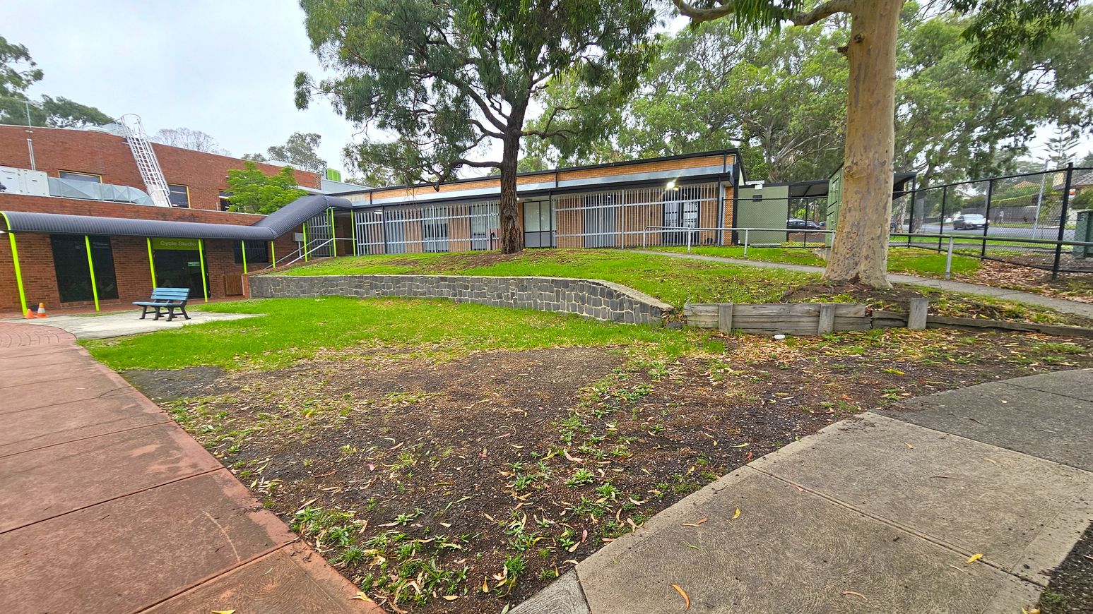 Outdoor grassed area. Functional and Barre Studio. Ivanhoe Aquatic Centre. A paved walkway leads to a brick building with large windows and metal bars. On the left, a blue bench sits on a concrete area near a "Cycle Studio" sign. A large tree stands in the centre, surrounded by grass and a low stone wall. The right side features a fenced area with more trees and a path. The ground is a mix of grass and bare patches, with scattered leaves. Outdoor grassed area. Functional and Barre Studio. Ivanhoe Aquatic Centre. A paved walkway leads to a brick building with large windows and metal bars. On the left, a blue bench sits on a concrete area near a "Cycle Studio" sign. A large tree stands in the centre, surrounded by grass and a low stone wall. The right side features a fenced area with more trees and a path. The ground is a mix of grass and bare patches, with scattered leaves.