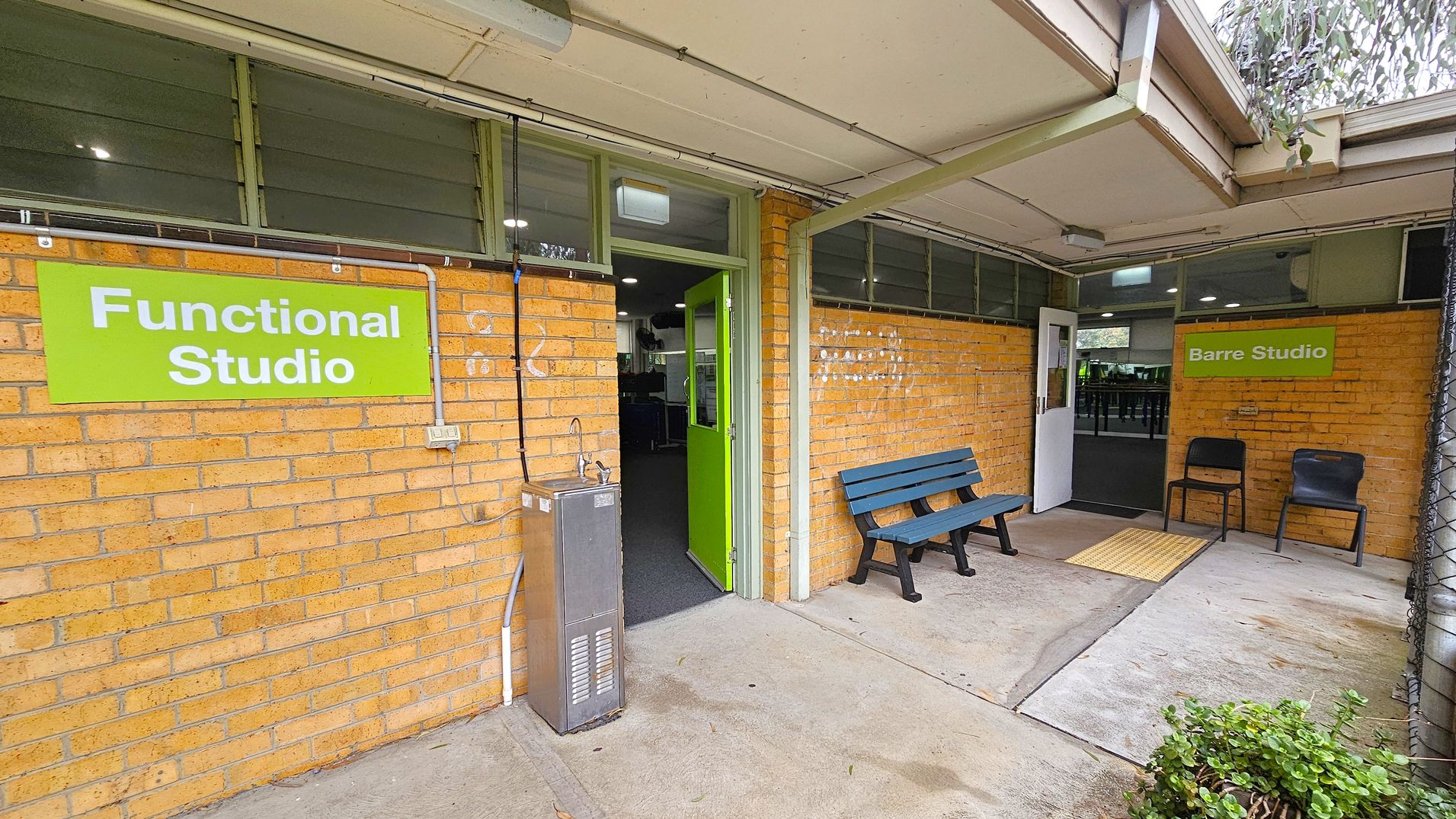 Studio doorway entrances. Functional and Barre Studio. Ivanhoe Aquatic Centre. A brick building with two entrances. On the left, a green sign reads "Functional Studio" above a green door, with a water fountain nearby. On the right, a sign says "Barre Studio" above a white door, with two chairs beside it. A blue bench is positioned between the entrances. The floor is concrete, and the ceiling is light-coloured with visible piping. A small bush is in the foreground near the right entrance. Studio doorway entrances. Functional and Barre Studio. Ivanhoe Aquatic Centre. A brick building with two entrances. On the left, a green sign reads "Functional Studio" above a green door, with a water fountain nearby. On the right, a sign says "Barre Studio" above a white door, with two chairs beside it. A blue bench is positioned between the entrances. The floor is concrete, and the ceiling is light-coloured with visible piping. A small bush is in the foreground near the right entrance.