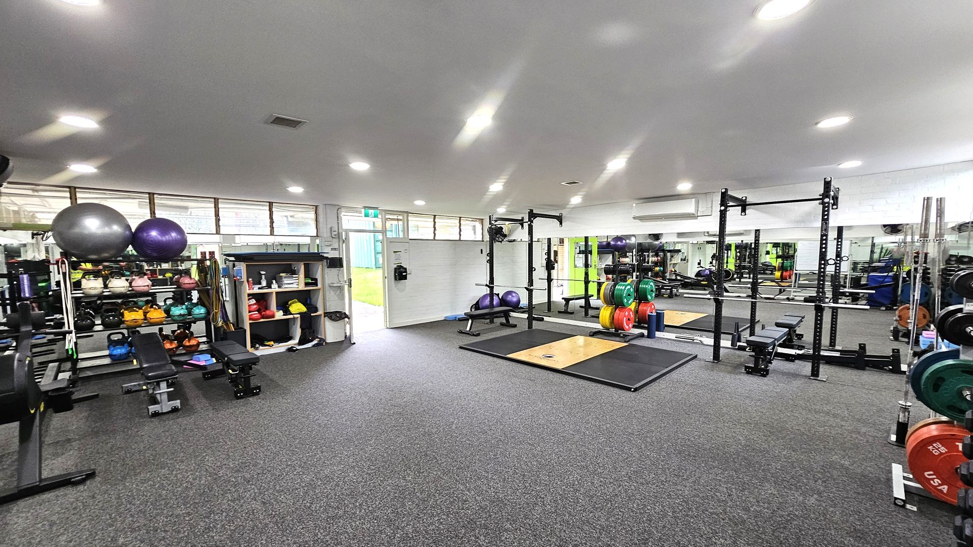 Functional studio and entry. Functional and Barre Studio. Ivanhoe Aquatic Centre. A gym with a grey carpeted floor and a white ceiling with recessed lights. On the left, there are shelves with kettlebells, medicine balls, and exercise balls. In the centre, a squat rack with colourful weight plates and a lifting platform is visible. The right side features more weightlifting equipment and mirrors along the wall. An open door at the back leads outside, allowing natural light to enter. Functional studio and entry. Functional and Barre Studio. Ivanhoe Aquatic Centre. A gym with a grey carpeted floor and a white ceiling with recessed lights. On the left, there are shelves with kettlebells, medicine balls, and exercise balls. In the centre, a squat rack with colourful weight plates and a lifting platform is visible. The right side features more weightlifting equipment and mirrors along the wall. An open door at the back leads outside, allowing natural light to enter.