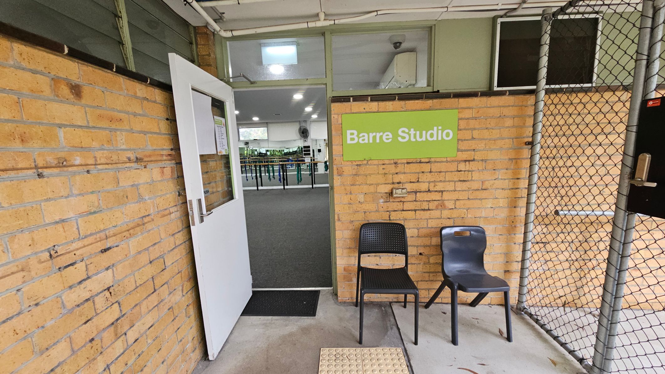 Barre Studio entrance. Functional and Barre Studio. Ivanhoe Aquatic Centre. A brick wall with a white door slightly open, leading into a room with a grey carpet and exercise equipment. A green sign on the wall reads "Barre Studio". Two chairs, one black and one grey, are placed outside the door. A chain-link fence is on the right side. The ceiling is white with visible pipes, and the floor is concrete with a textured mat near the entrance. Barre Studio entrance. Functional and Barre Studio. Ivanhoe Aquatic Centre. A brick wall with a white door slightly open, leading into a room with a grey carpet and exercise equipment. A green sign on the wall reads "Barre Studio". Two chairs, one black and one grey, are placed outside the door. A chain-link fence is on the right side. The ceiling is white with visible pipes, and the floor is concrete with a textured mat near the entrance.