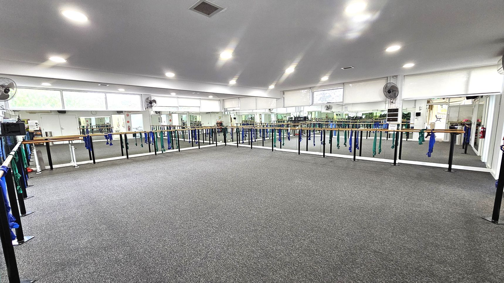 Mirrored walls Barre Studio. Functional and Barre Studio. Ivanhoe Aquatic Centre. A spacious dance studio with grey carpeted flooring. The room features mirrored walls on the left and right, with ballet barres running along them. Blue and green resistance bands hang from the barres. The ceiling is white with recessed lighting, and there are several wall-mounted fans. Large windows above the mirrors allow natural light to enter. The room is empty, providing ample space for movement. Mirrored walls Barre Studio. Functional and Barre Studio. Ivanhoe Aquatic Centre. A spacious dance studio with grey carpeted flooring. The room features mirrored walls on the left and right, with ballet barres running along them. Blue and green resistance bands hang from the barres. The ceiling is white with recessed lighting, and there are several wall-mounted fans. Large windows above the mirrors allow natural light to enter. The room is empty, providing ample space for movement.