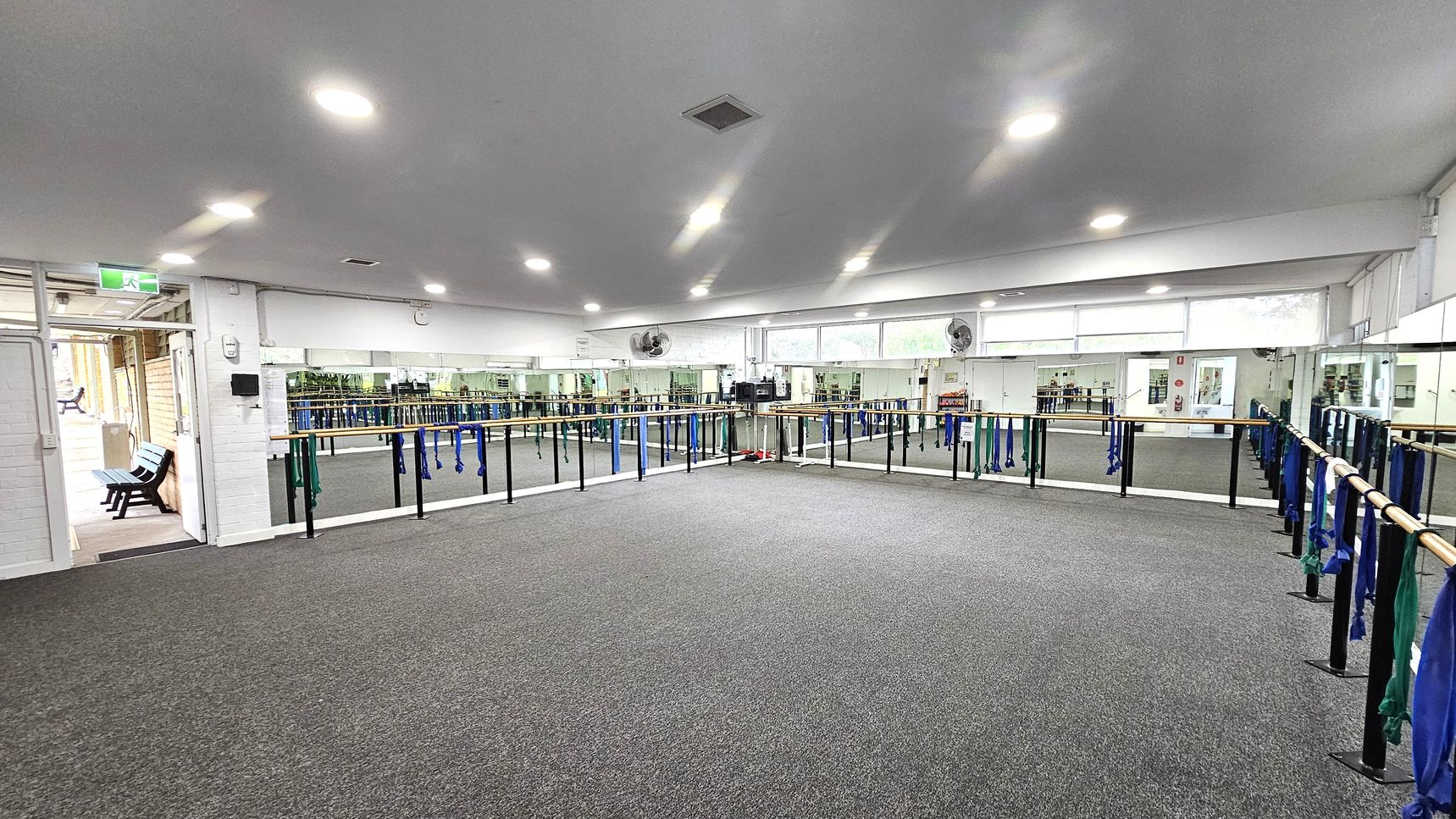 Mirrored walls and doorway Barre Studio. Functional and Barre Studio. Ivanhoe Aquatic Centre. A spacious dance studio with grey carpeted flooring and a white ceiling with recessed lights. The walls are lined with mirrors, and ballet barres run along them, adorned with blue and green resistance bands. On the left, there's an open door leading to a hallway with a bench. The room is well-lit, and there are ceiling fans above the mirrors. Mirrored walls and doorway Barre Studio. Functional and Barre Studio. Ivanhoe Aquatic Centre. A spacious dance studio with grey carpeted flooring and a white ceiling with recessed lights. The walls are lined with mirrors, and ballet barres run along them, adorned with blue and green resistance bands. On the left, there's an open door leading to a hallway with a bench. The room is well-lit, and there are ceiling fans above the mirrors.