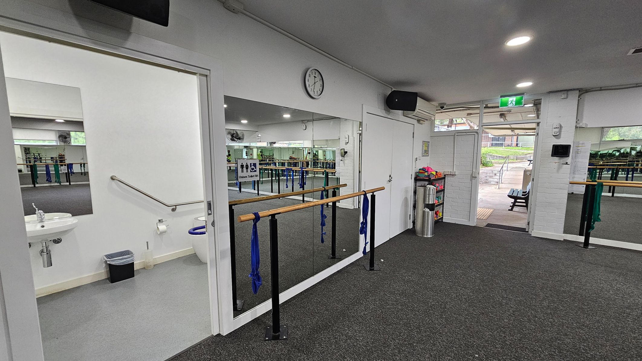 Accessible bathroom wayfinding Barre Studio. Functional and Barre Studio. Ivanhoe Aquatic Centre. A dance studio with a grey carpeted floor. On the left, there's an open door leading to a bathroom with a sink, toilet, and handrail. In the centre, a mirrored wall with a wooden ballet barre attached, adorned with blue fabric. A clock is mounted above the mirror. On the right, an open door leads outside, showing a paved area with benches. A shelf with colourful items and a bin is near the door. Accessible bathroom wayfinding Barre Studio. Functional and Barre Studio. Ivanhoe Aquatic Centre. A dance studio with a grey carpeted floor. On the left, there's an open door leading to a bathroom with a sink, toilet, and handrail. In the centre, a mirrored wall with a wooden ballet barre attached, adorned with blue fabric. A clock is mounted above the mirror. On the right, an open door leads outside, showing a paved area with benches. A shelf with colourful items and a bin is near the door.