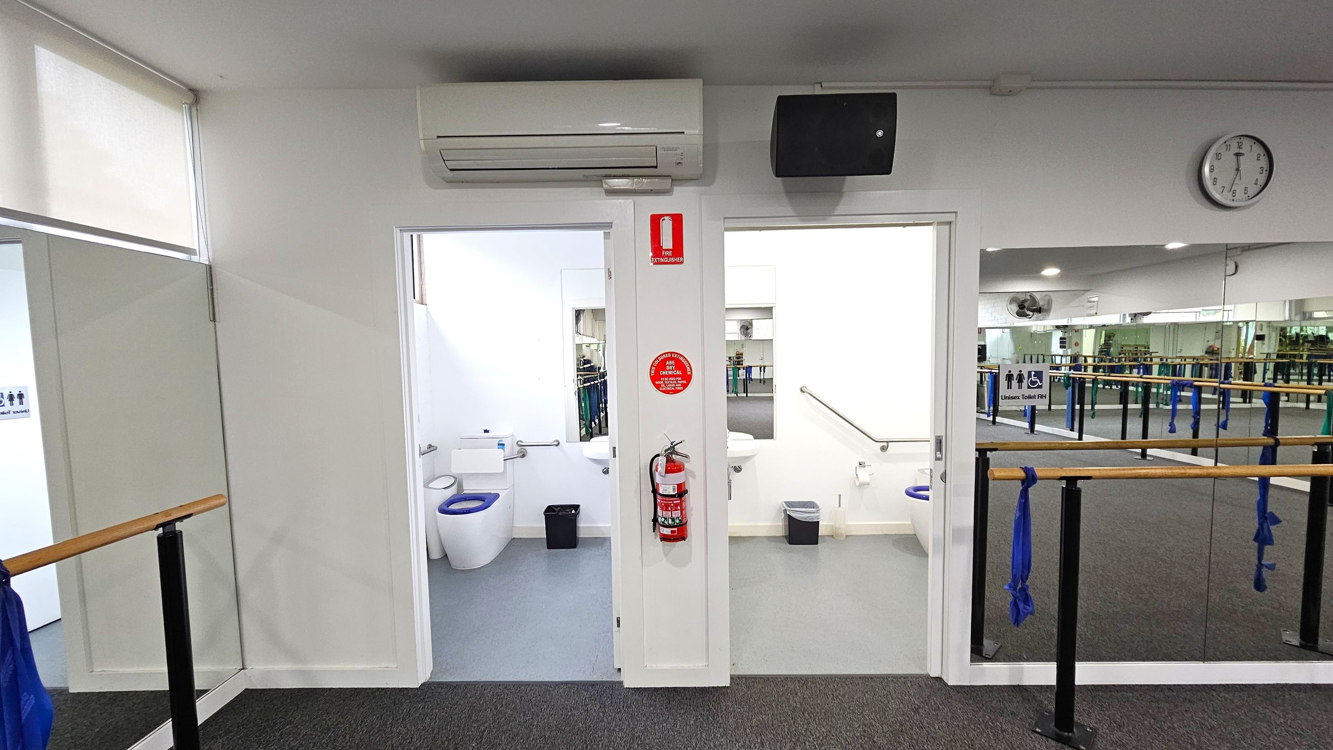 Two accessible bathrooms. Functional and Barre Studio. Ivanhoe Aquatic Centre. A room with two open doors leading to toilets, each with a white toilet and a small bin. Above the doors, an air conditioning unit and a speaker are mounted. A fire extinguisher is attached to the wall between the doors. On the right, a wall clock shows the time. The foreground features a mirrored wall with ballet barres and blue fabric tied to them. The floor is carpeted, and the ceiling has recessed lighting. Two accessible bathrooms. Functional and Barre Studio. Ivanhoe Aquatic Centre. A room with two open doors leading to toilets, each with a white toilet and a small bin. Above the doors, an air conditioning unit and a speaker are mounted. A fire extinguisher is attached to the wall between the doors. On the right, a wall clock shows the time. The foreground features a mirrored wall with ballet barres and blue fabric tied to them. The floor is carpeted, and the ceiling has recessed lighting.