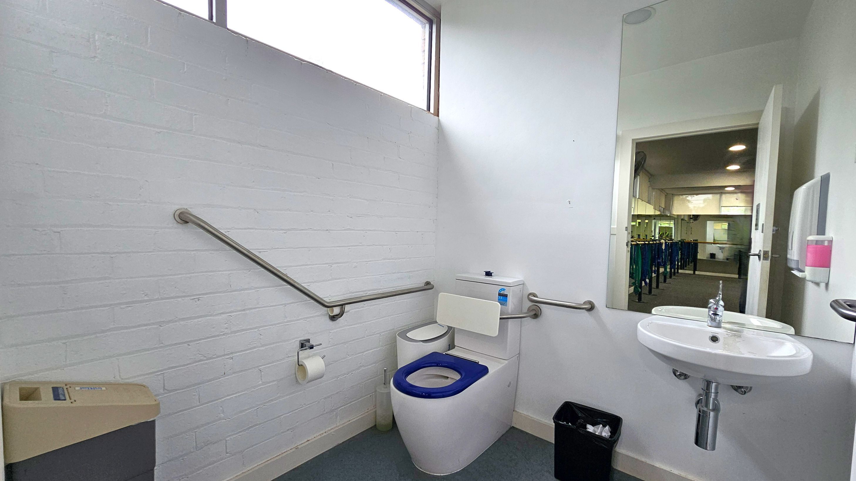 Internal aspect accessible bathroom. Functional and Barre Studio. Ivanhoe Aquatic Centre. A bathroom with white walls and a grey floor. On the left, there is a beige bin and a metal handrail along the wall. In the centre, a toilet with a blue seat and a white backrest is positioned against the wall, with a toilet roll holder nearby. On the right, a white sink with a chrome tap is mounted below a large mirror reflecting a room with tables and chairs. A soap dispenser is mounted next to the sink. Internal aspect accessible bathroom. Functional and Barre Studio. Ivanhoe Aquatic Centre. A bathroom with white walls and a grey floor. On the left, there is a beige bin and a metal handrail along the wall. In the centre, a toilet with a blue seat and a white backrest is positioned against the wall, with a toilet roll holder nearby. On the right, a white sink with a chrome tap is mounted below a large mirror reflecting a room with tables and chairs. A soap dispenser is mounted next to the sink.