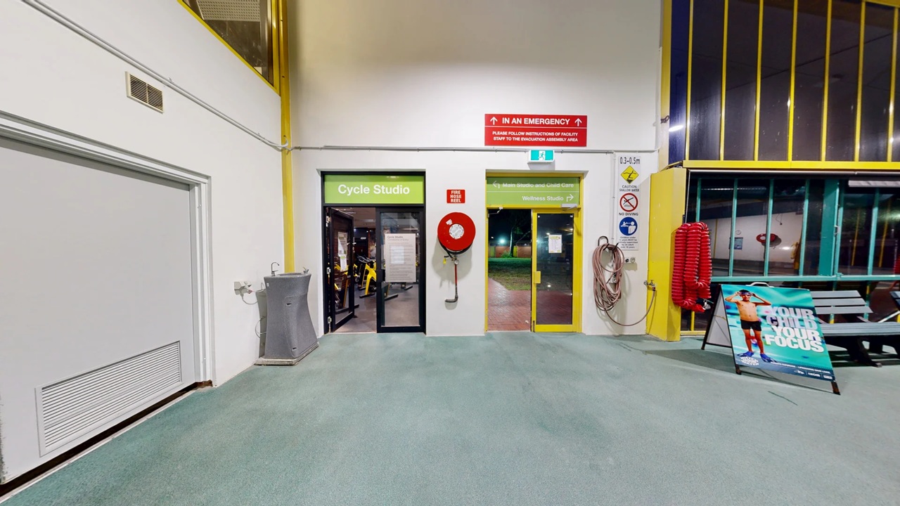 A hallway with a green carpeted floor. On the left, there's a water fountain and a door labelled "Cycle Studio" with exercise equipment visible inside. In the centre, a yellow-framed glass door leads outside, with a fire hose reel mounted on the wall. On the right, a coiled hose and a sign with safety instructions are visible. A poster stand with an image of a person is near a bench. The walls are white with yellow accents, and the ceiling is not visible. A hallway with a green carpeted floor. On the left, there's a water fountain and a door labelled "Cycle Studio" with exercise equipment visible inside. In the centre, a yellow-framed glass door leads outside, with a fire hose reel mounted on the wall. On the right, a coiled hose and a sign with safety instructions are visible. A poster stand with an image of a person is near a bench. The walls are white with yellow accents, and the ceiling is not visible.