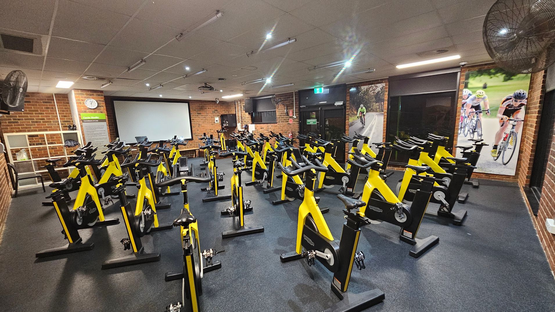 A cycling studio with numerous yellow stationary bikes arranged in rows on a dark floor. The walls are brick, with large posters of cyclists on the right. At the back, there's a large screen and a clock on the left wall. The ceiling is fitted with lights and fans. A cycling studio with numerous yellow stationary bikes arranged in rows on a dark floor. The walls are brick, with large posters of cyclists on the right. At the back, there's a large screen and a clock on the left wall. The ceiling is fitted with lights and fans.