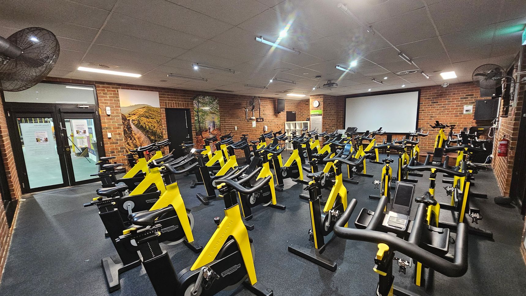 A gym room with numerous yellow and black stationary bikes arranged in rows on a dark floor. The walls are brick, with a large screen at the front. On the left, there are glass doors with notices and a wall mural depicting a landscape. Ceiling fans and lights are visible above. A gym room with numerous yellow and black stationary bikes arranged in rows on a dark floor. The walls are brick, with a large screen at the front. On the left, there are glass doors with notices and a wall mural depicting a landscape. Ceiling fans and lights are visible above.
