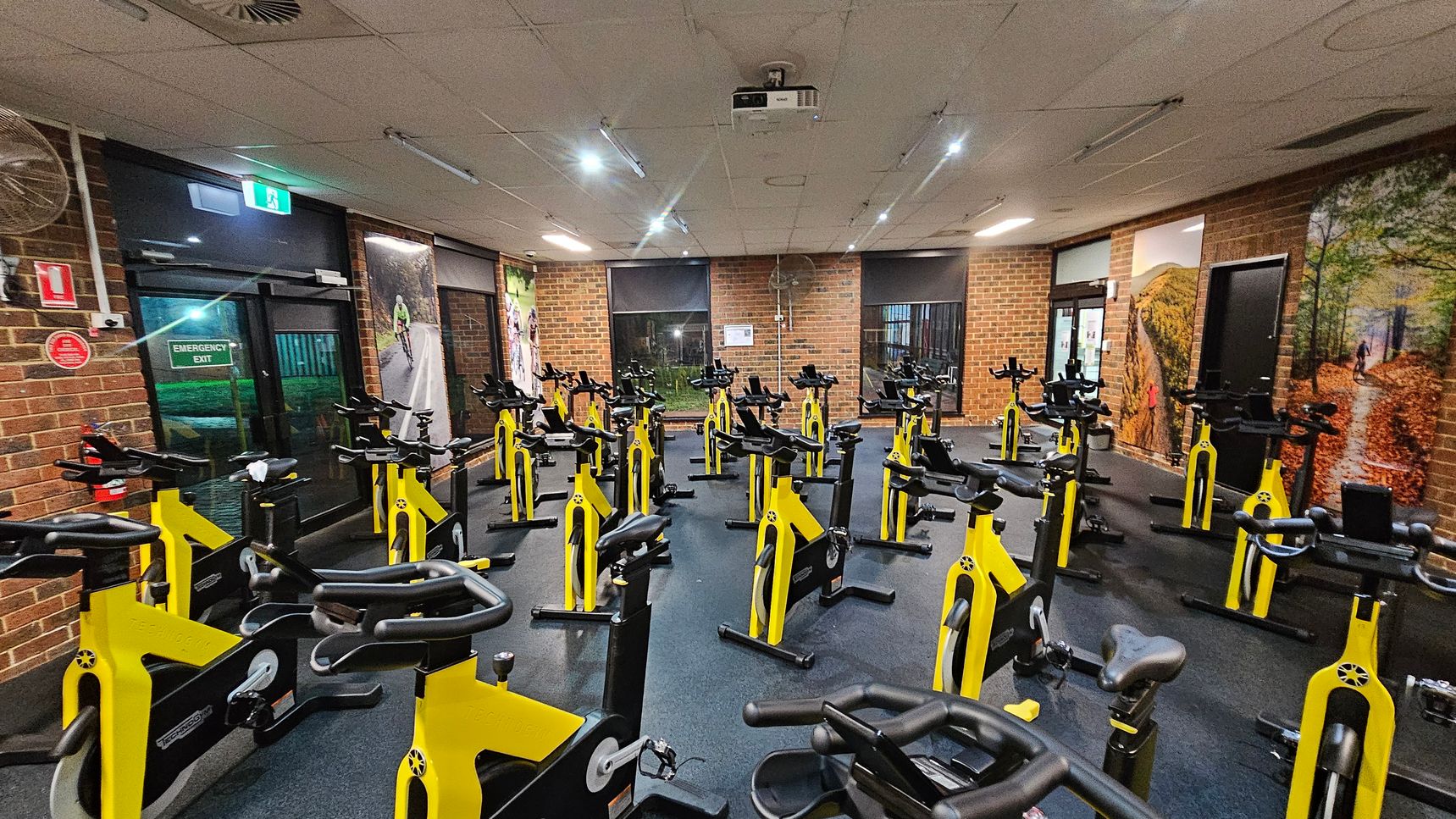 A room with numerous yellow stationary bikes arranged in rows on a dark floor. The walls are brick, featuring large cycling-themed murals. On the left, there's an emergency exit door with a green sign above it. The ceiling is fitted with fluorescent lights and a projector. The room is well-lit, and there are windows with blinds on the far wall. A room with numerous yellow stationary bikes arranged in rows on a dark floor. The walls are brick, featuring large cycling-themed murals. On the left, there's an emergency exit door with a green sign above it. The ceiling is fitted with fluorescent lights and a projector. The room is well-lit, and there are windows with blinds on the far wall.