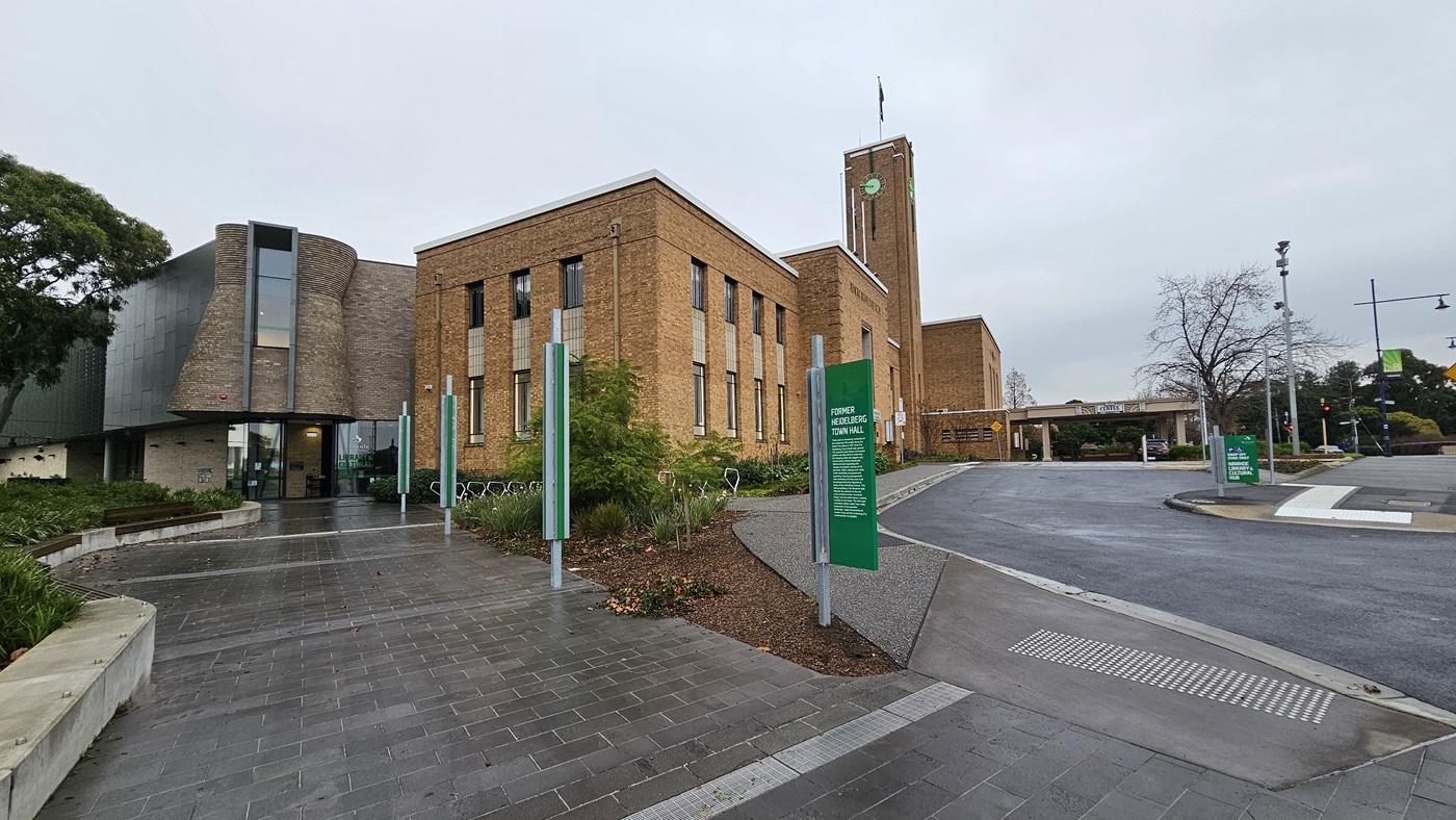 On the left, there is a modern building with a curved glass facade and a garden with green shrubs and trees. In the middle, there are several green signs with white text and a pathway made of grey tiles. On the right, a large brick building with tall windows and a clock tower. The foreground features a grey tiled floor and a wet asphalt road with a zebra crossing.