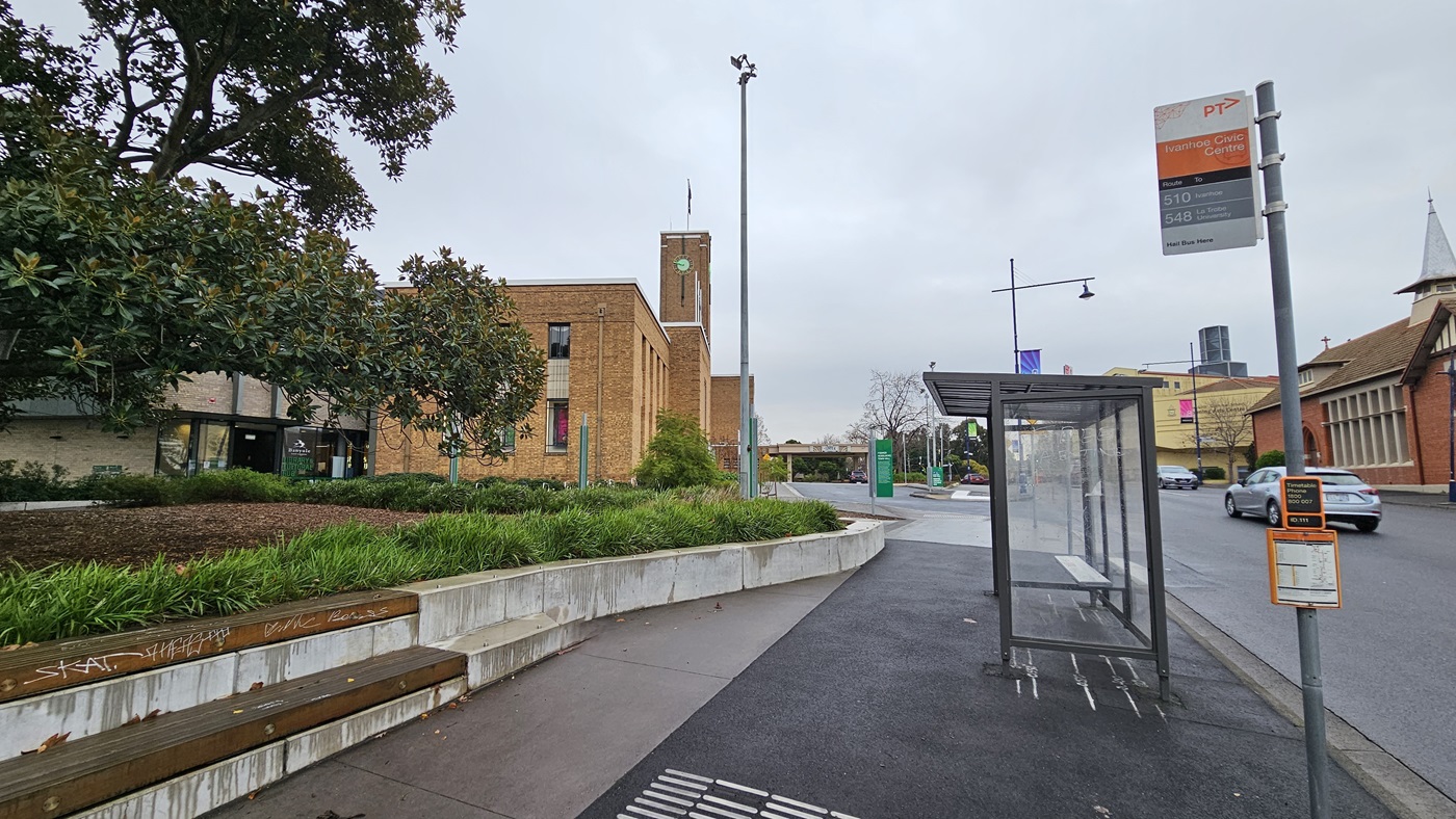 On the left, there is a large tree with dense foliage and a garden area with green shrubs and a curved concrete bench. In the middle, a brick building with a clock tower and a flagpole. On the right, a bus stop with a transparent shelter and a sign displaying bus routes. The foreground features a wet asphalt road and a concrete pavement. The background includes additional buildings and streetlights.