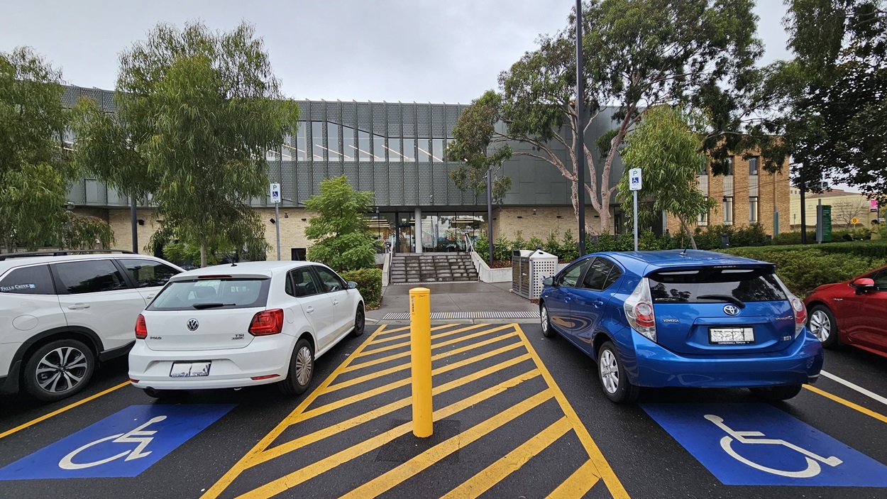 On the left, there is a white car parked in a disabled parking space next to a grey SUV. In the middle, a building with a modern grey facade and large windows, with a set of stairs leading to the entrance. On the right, a blue car parked in another disabled parking space. The foreground features yellow and black striped markings on the asphalt, indicating the parking area. Trees and shrubs are visible around the parking lot.