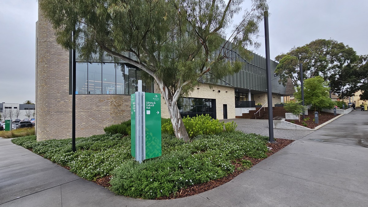 On the left, there is a green sign with white text in front of a tree surrounded by a garden with green shrubs and white flowers. In the middle, a modern building with a brick facade and large windows. On the right, a pathway with steps leading up to the building entrance, bordered by more greenery and trees. The foreground features a grey concrete pavement, and the background includes additional trees and parts of other buildings.