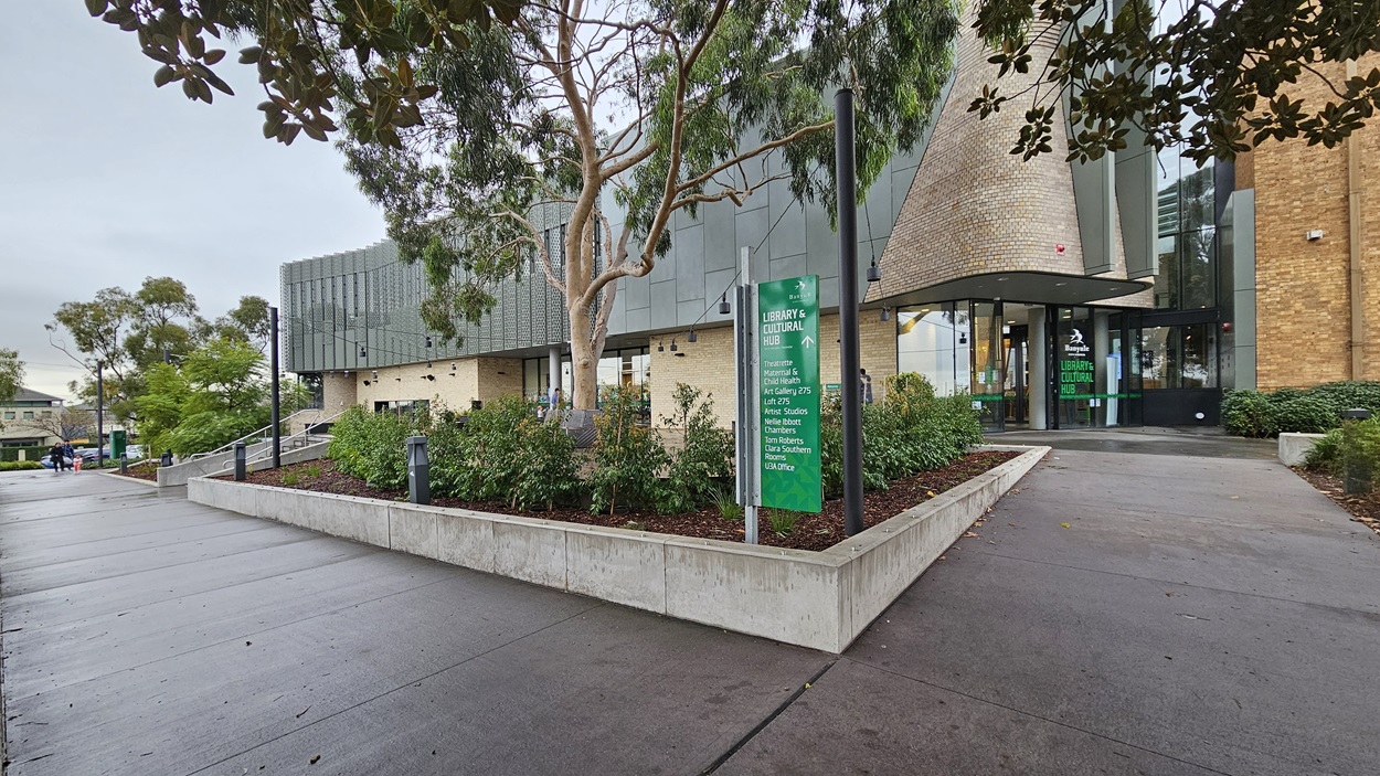 On the left, there is a concrete pathway bordered by a low concrete wall with green shrubs and a tall tree. In the middle, a modern building with a grey facade, large windows, and a cylindrical brick structure near the entrance. On the right, another concrete pathway leads to the building entrance, flanked by more greenery and a green sign with white text. The floor is made of smooth grey concrete.