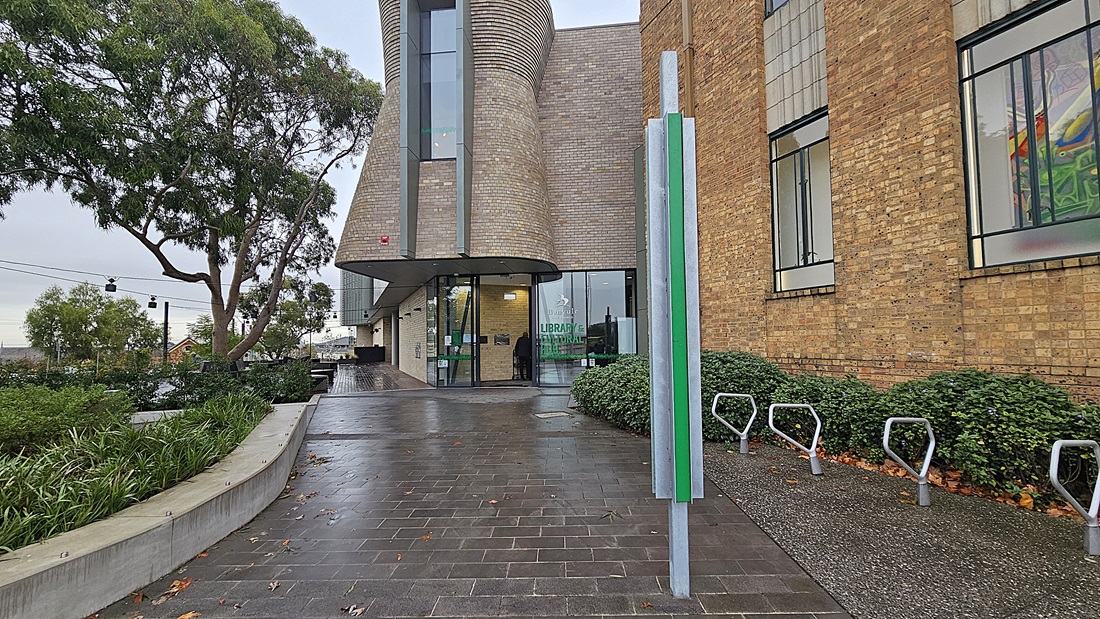 On the left, there is a curved concrete bench with green shrubs and a tall tree behind it. In the middle, a modern building with a cylindrical brick structure and large glass doors leading to the entrance. On the right, the building continues with a brick facade and tall windows. The foreground features a wet, dark grey tiled floor, and there are bike racks and a green sign with white text near the entrance.