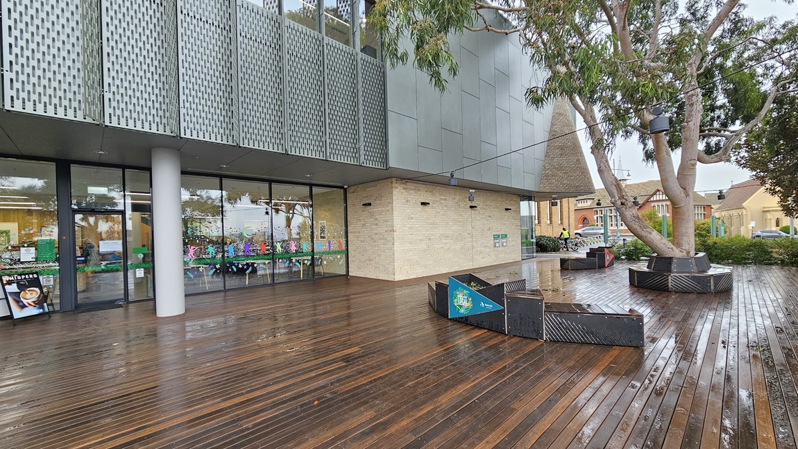 On the left, there is a modern building with large glass windows and a perforated metal facade above. In the middle, the building's entrance with glass doors and a brick wall section. On the right, there are large planters with trees and a few benches. The foreground features a wet wooden deck floor. The background includes additional buildings and greenery.