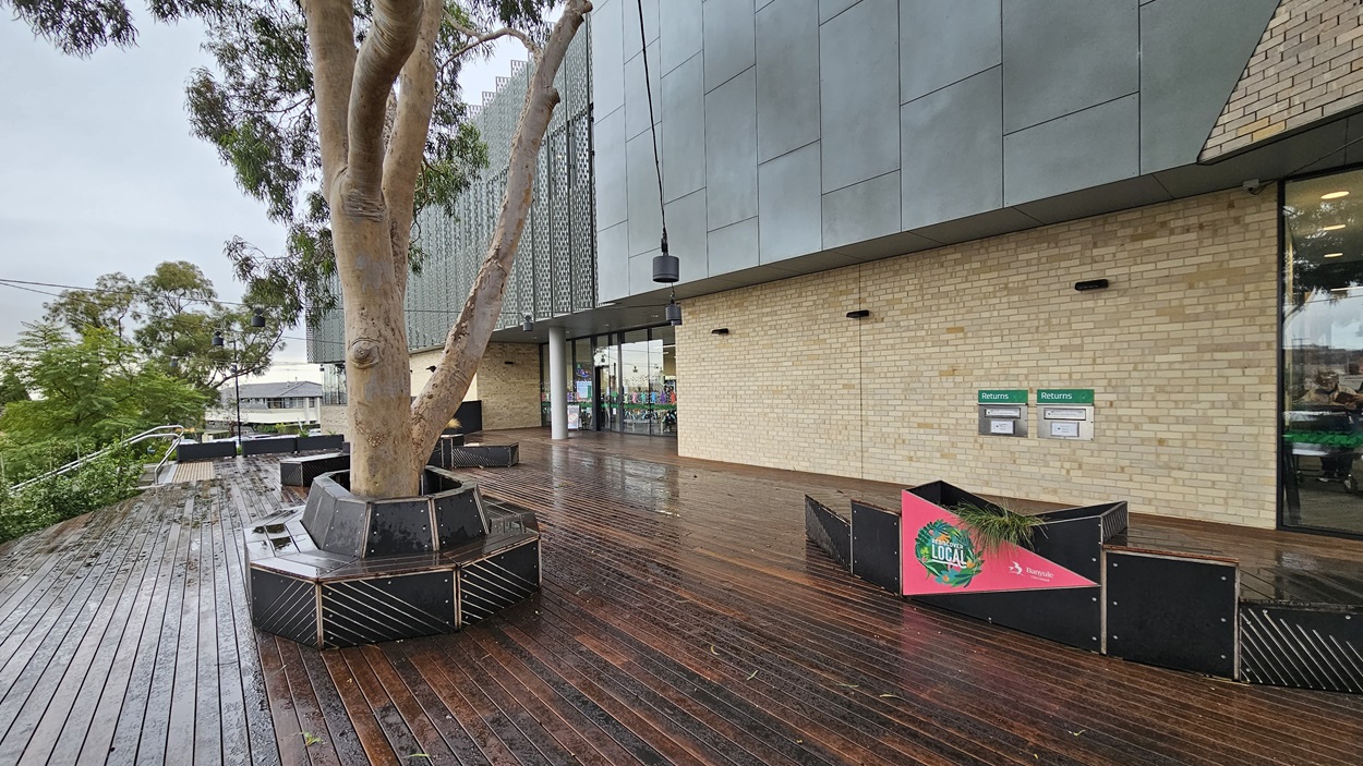 On the left, there is a large tree planted in a black, rectangular planter on a wooden deck. In the middle, the entrance to a modern building with glass doors and a brick wall. On the right, there are two green signs with white text above a black planter containing shrubs. The floor is made of wet wooden planks, and the ceiling is a smooth grey surface with recessed lighting. The background includes more trees and parts of other buildings.