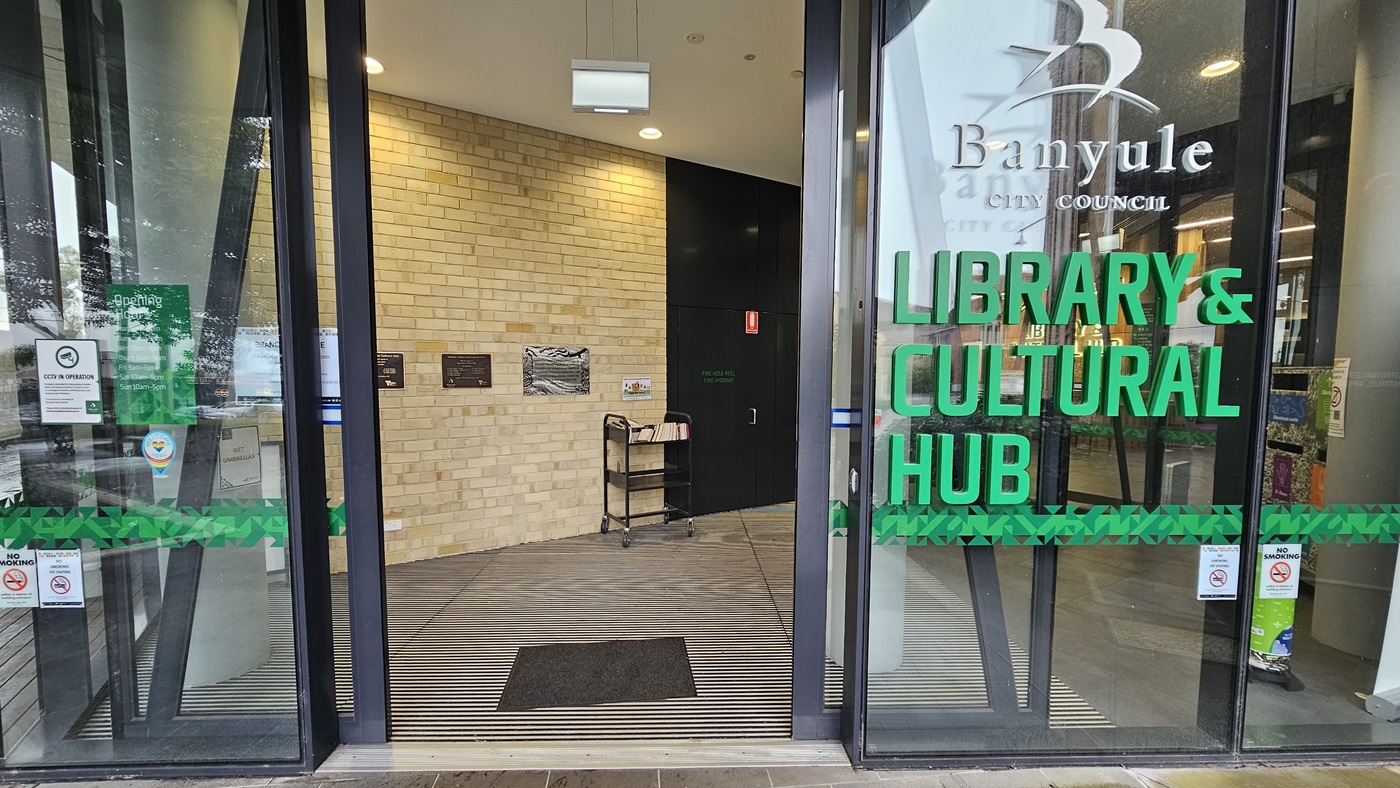 On the left, there is a glass door with a green stripe and various stickers. In the middle, a brick wall with a small table holding brochures and a black mat on the floor. On the right, another glass door with the text "Banyule City Council" and "Library & Cultural Hub" in green. The floor is a combination of wooden planks and a ribbed mat at the entrance. The ceiling is white with recessed lighting and a hanging light fixture.