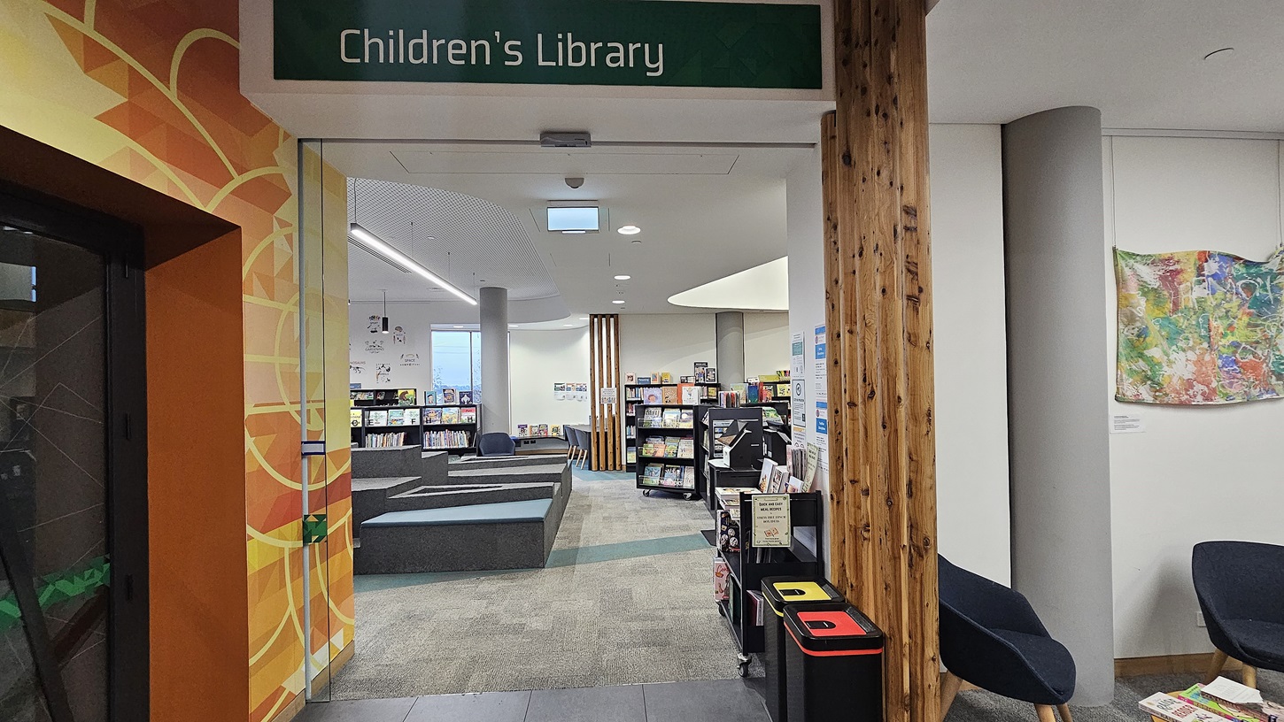 On the left, there is a colourful mural with geometric patterns and a glass door. In the middle, a sign reading "Children's Library" hangs above the entrance, leading to a room with tiered seating and bookshelves filled with books. On the right, there is a wooden column, a recycling station with bins, and a grey armchair next to a cylindrical pillar. The floor is a mix of grey tiles and carpet, and the ceiling is white with recessed lighting and a skylight.