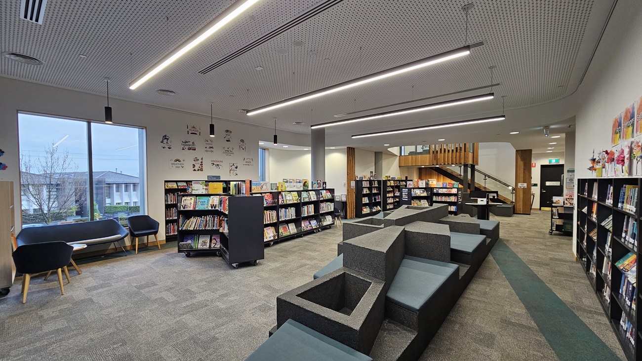 On the left, there are large windows with two black armchairs and a small table in front of them. In the middle, there are several bookshelves filled with books and a tiered seating area with grey cushions. On the right, more bookshelves line the wall, and a wooden staircase with a glass railing leads to an upper level. The floor is covered in grey carpet tiles, and the ceiling is white with a perforated design and linear recessed lighting.