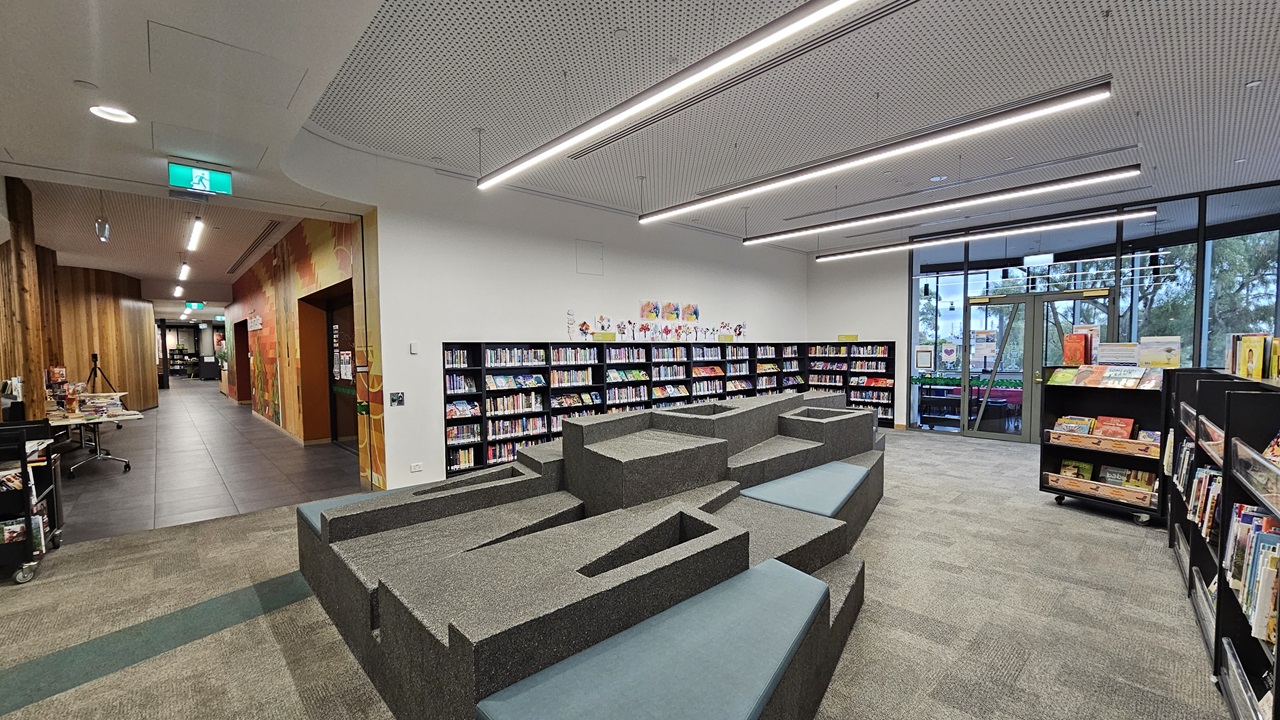 On the left, there is a hallway with wooden walls and a green exit sign above the door. In the middle, there is a grey, tiered seating area with blue cushions and integrated steps. On the right, there are bookshelves filled with books and a large window with a view of trees and buildings outside. The floor is covered in grey carpet tiles with a green stripe, and the ceiling is white with a perforated design and linear recessed lighting.