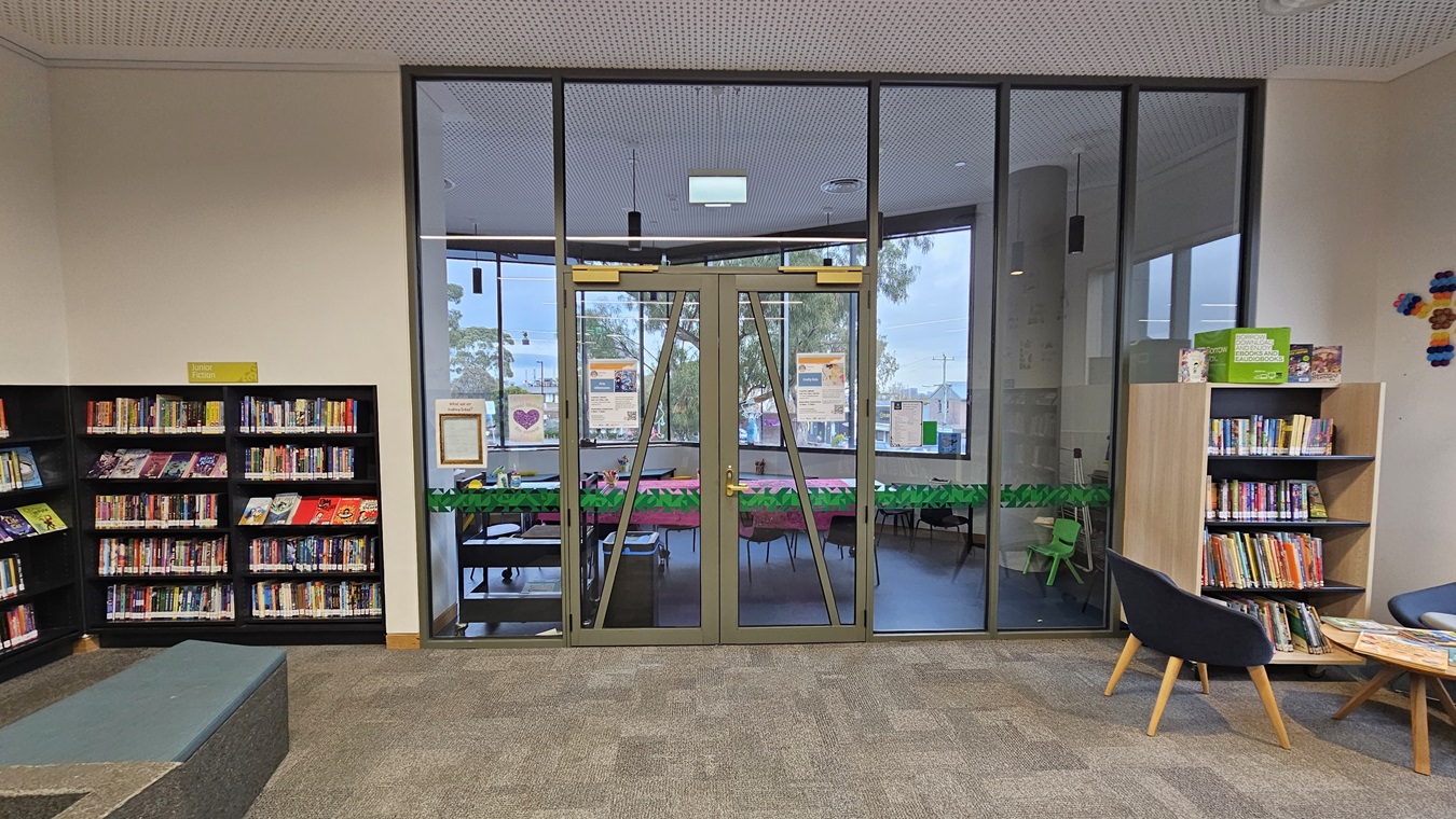 On the left, there are black bookshelves filled with books and a grey, tiered seating area with blue cushions. In the middle, there is a large glass door with metal frames leading to a room with tables and green chairs. On the right, there is a wooden bookshelf with books and a black armchair with a small round table beside it. The floor is covered in grey carpet tiles, and the ceiling is white with a perforated design and recessed lighting.