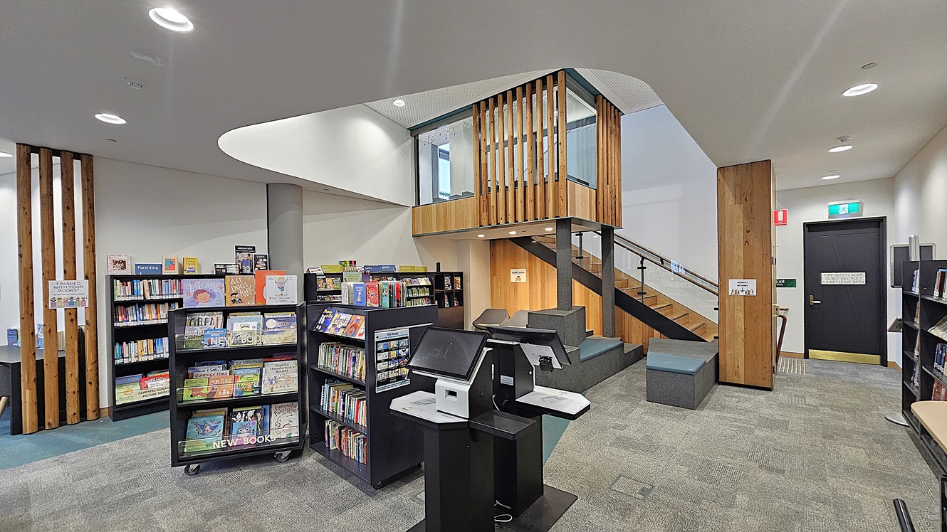 On the left, there are black bookshelves filled with books and a grey column. In the middle, there is a staircase with wooden slats and a glass railing leading to an upper level, with grey seating and a wooden wall behind it. On the right, there is a black door with a green exit sign above it and a fire extinguisher mounted on the wooden wall. The floor is covered in grey carpet tiles, and the ceiling is white with recessed lighting.