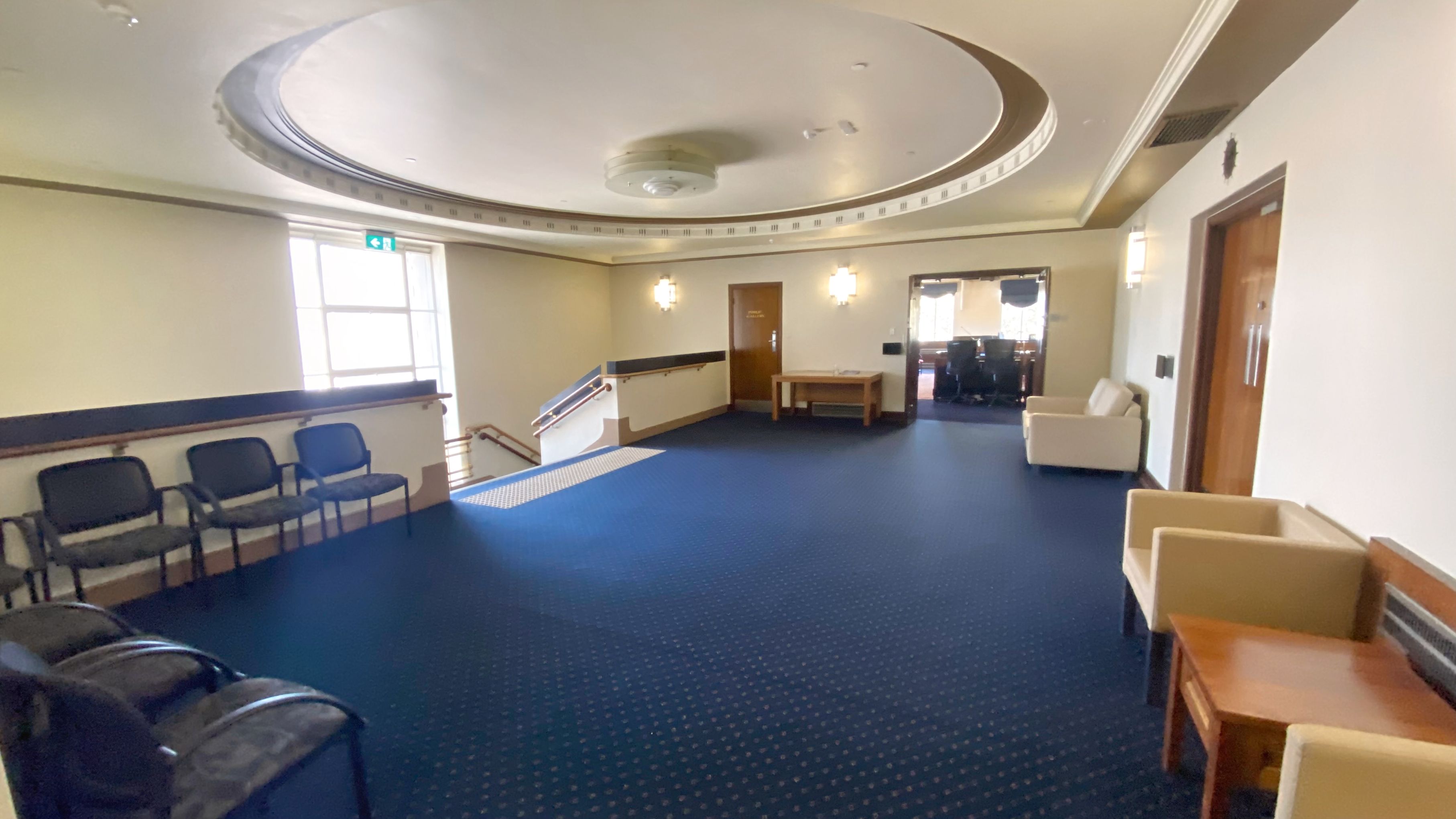 A spacious room with a blue carpet and cream walls. On the left, there are several black chairs lined up against the wall near a window with an exit sign. In the centre, a staircase with a handrail leads downwards. On the right, there are beige armchairs and a wooden table. Further back, a door and a small table are visible, with another room in the background. The ceiling features a large circular design with a central light fixture.