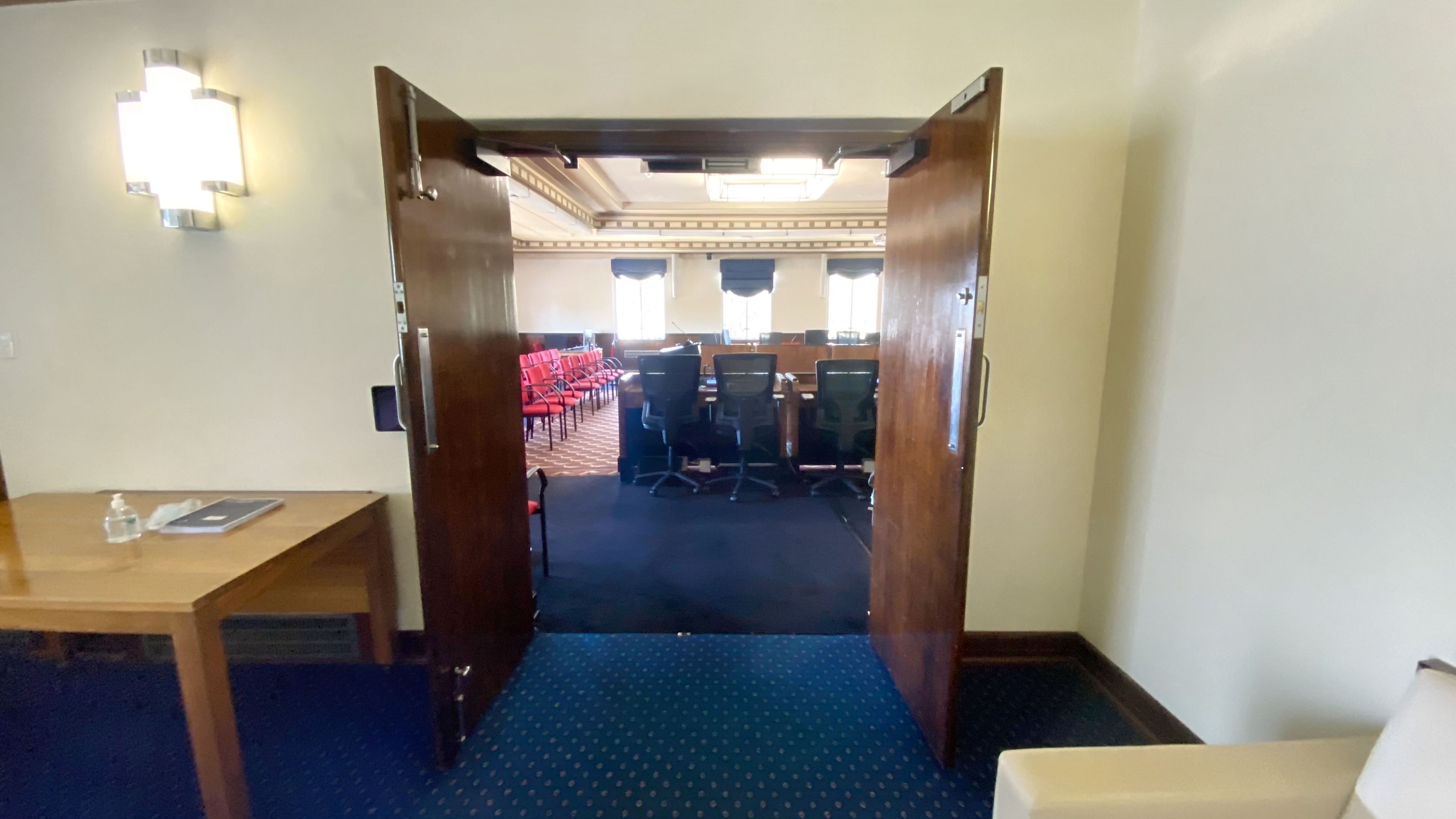 A room with wooden double doors open, leading to a larger room with rows of red chairs and black office chairs facing a desk. The floor is carpeted in blue with a dotted pattern. On the left, a wooden table holds a water bottle and a folder. A wall-mounted light fixture is above the table. The ceiling has decorative moulding and a large light fixture.