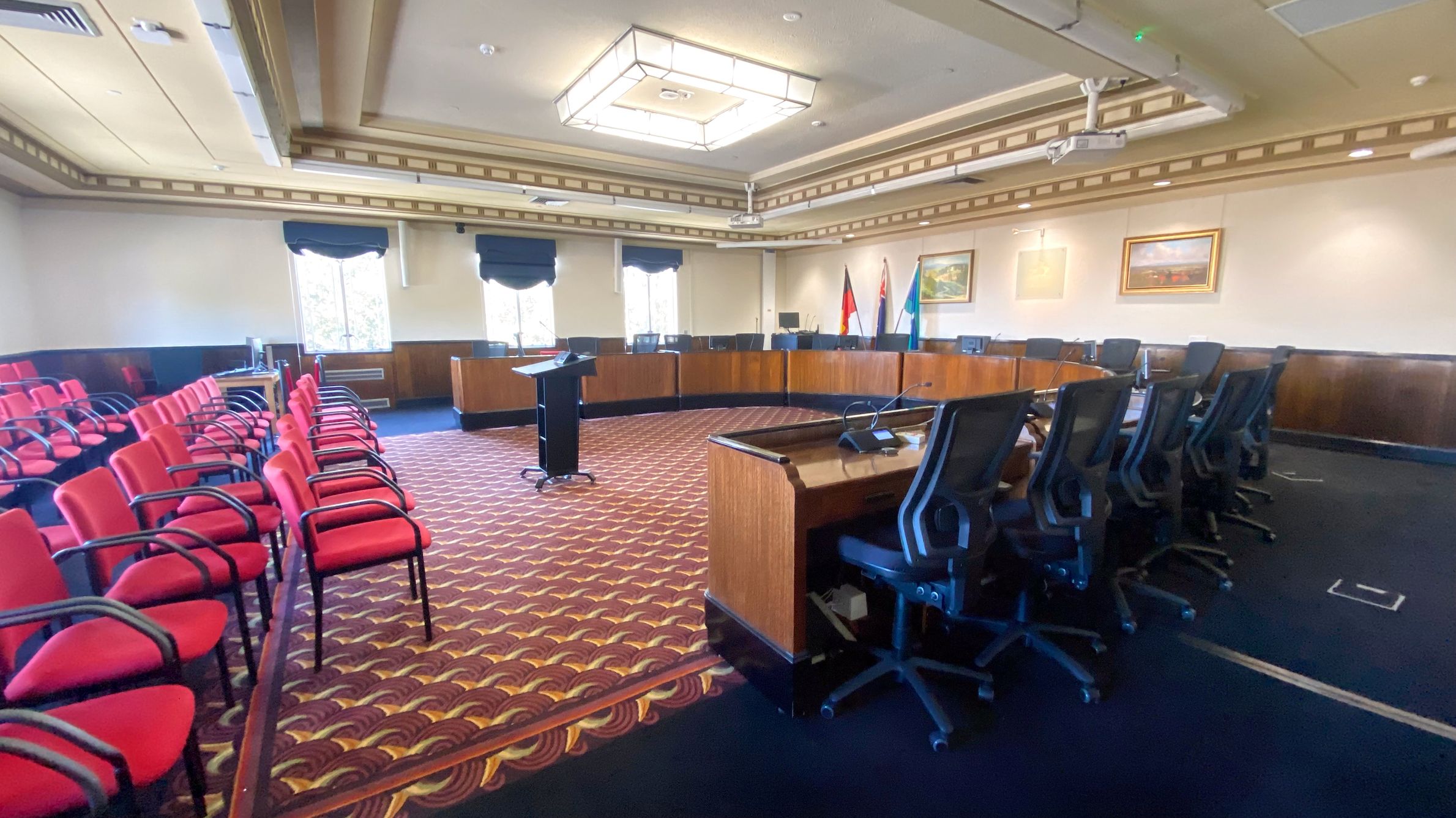 A conference room with a U-shaped arrangement of black office chairs and wooden desks on the right. In the foreground, a podium stands on a patterned carpet. On the left, rows of red chairs with black armrests face the centre. The walls are adorned with framed pictures and flags, and there are windows with dark curtains. The ceiling features a large, square light fixture.
