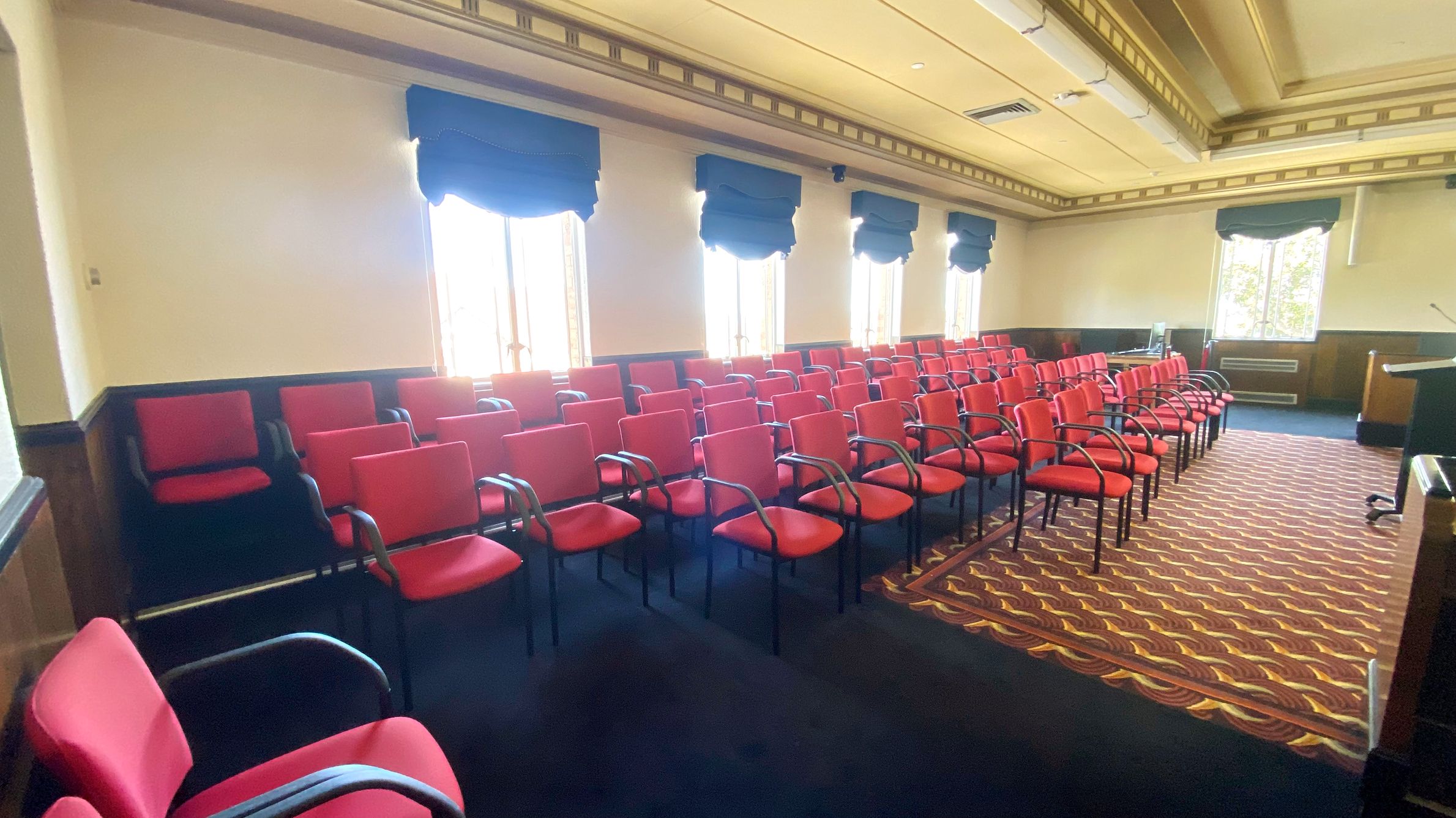 A room with rows of red chairs with black armrests arranged in a grid pattern. Large windows with dark blue curtains line the left wall, allowing natural light to enter. The floor is covered with a patterned carpet in shades of brown and gold. The ceiling is ornate with decorative moulding and recessed lighting. A podium is visible on the right side near the front of the room.